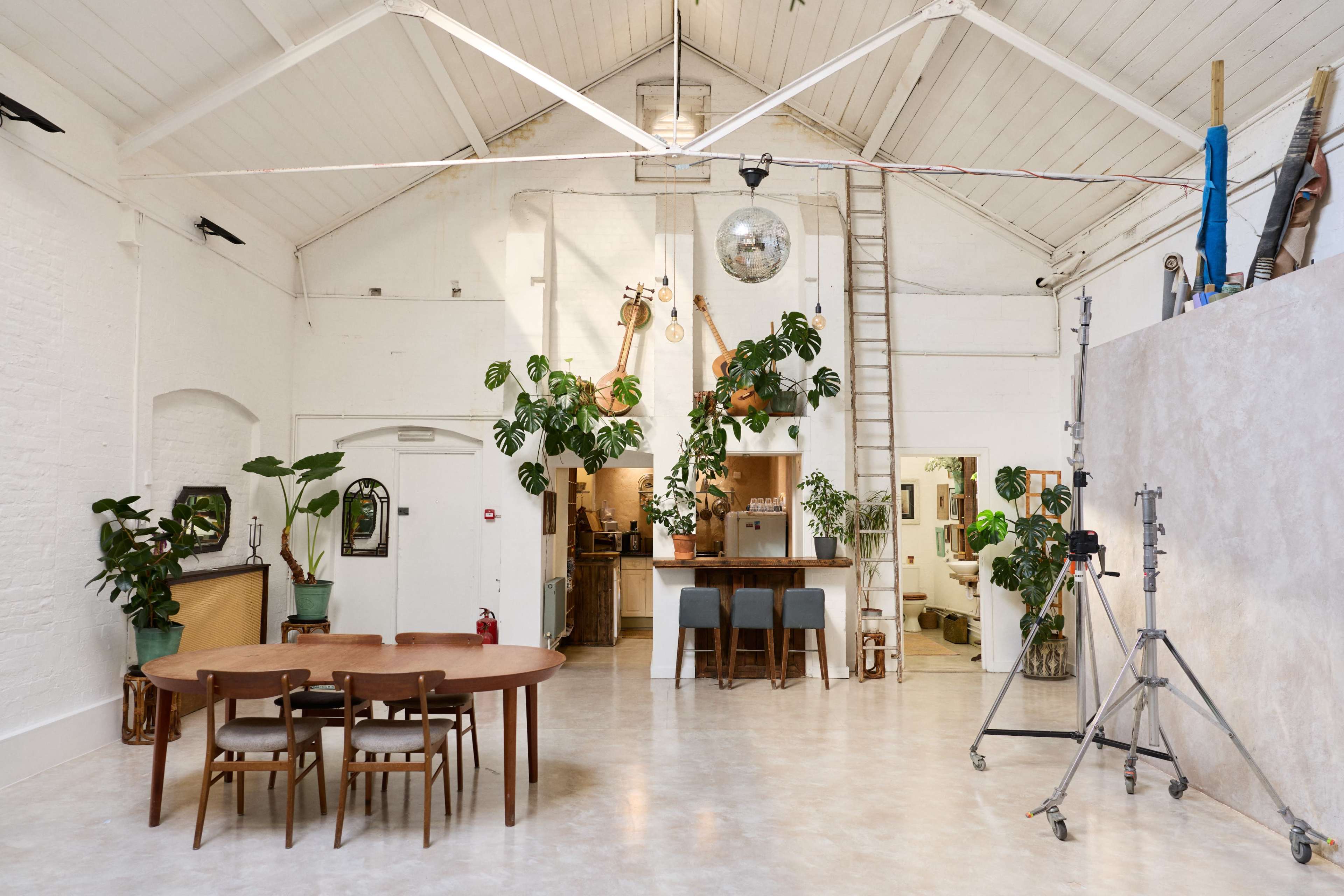 The image shows a spacious, well-lit studio with a dining table and chairs, a kitchen area in the background, and various plants placed throughout.