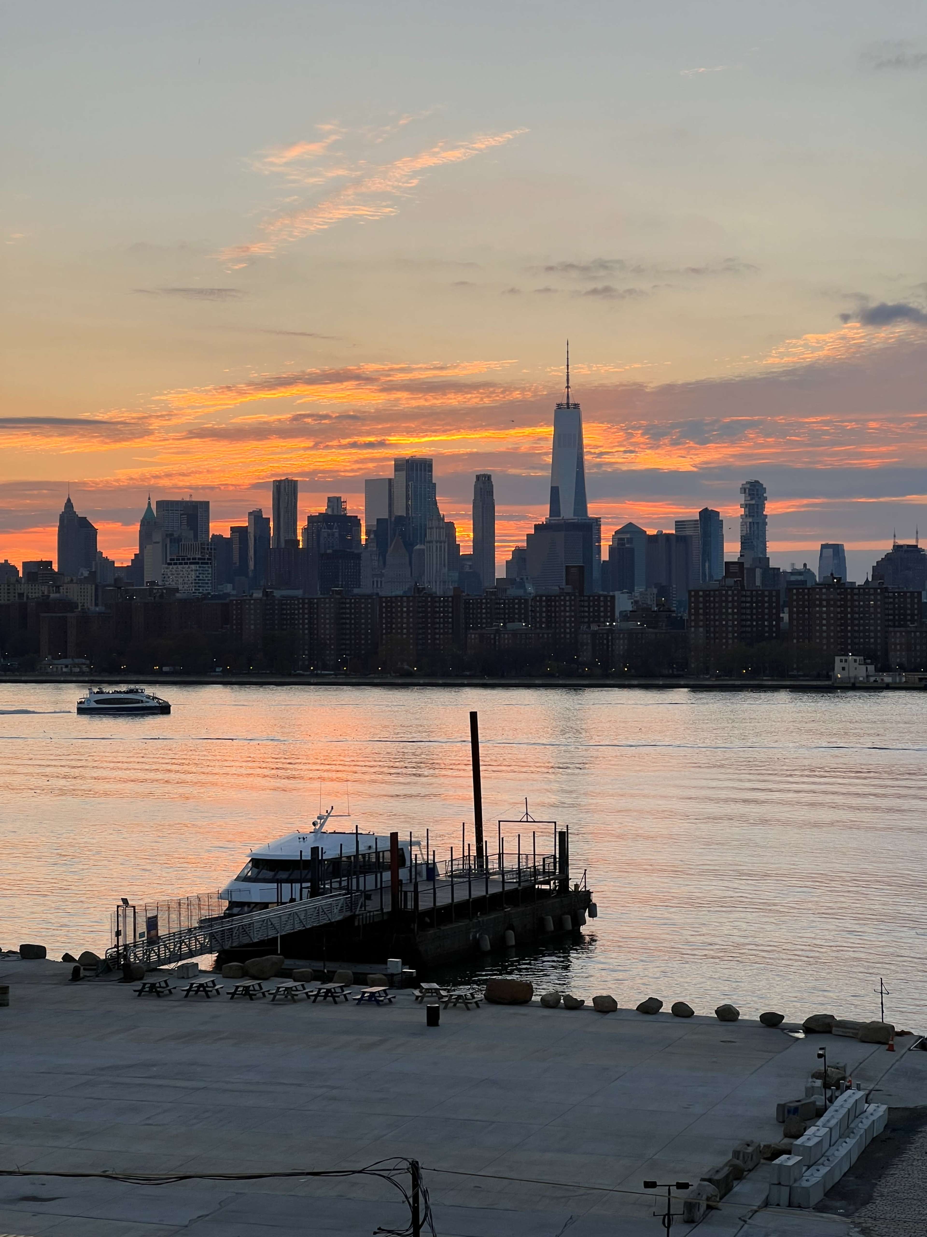 The image shows a view of the New York City skyline at sunset, with the One World Trade Center prominent and a boat docked at the waterfront.