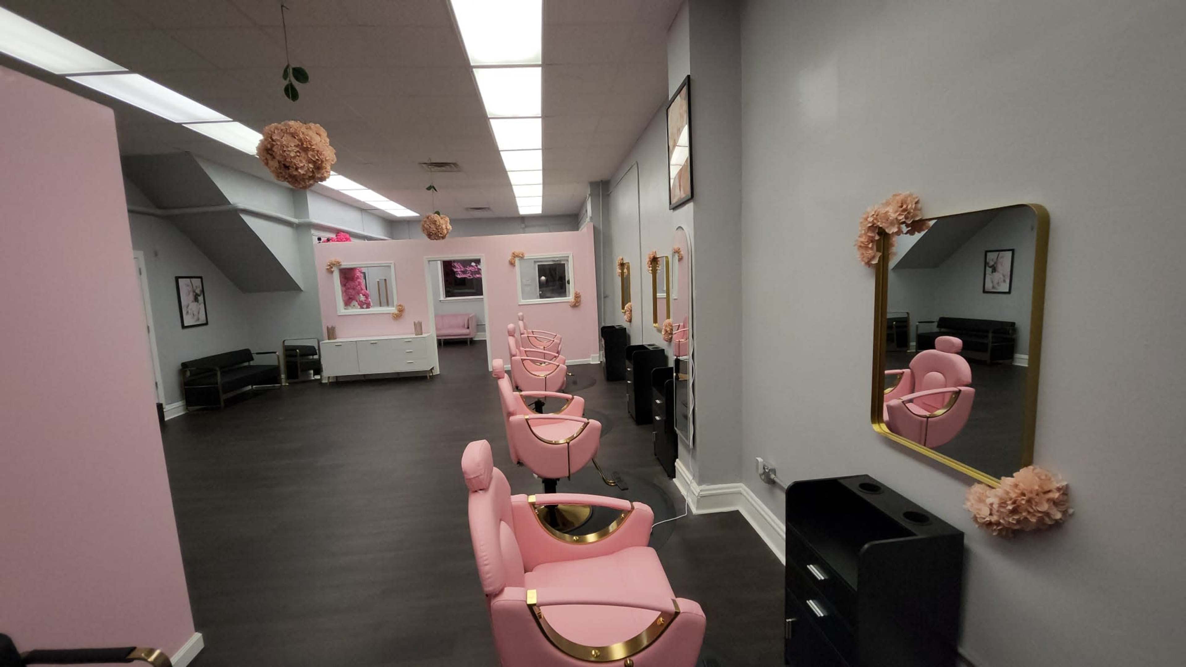 The image shows a hair salon with pink and gray walls, featuring several pink salon chairs arranged in a spacious layout, with decorative elements hanging from the ceiling.
