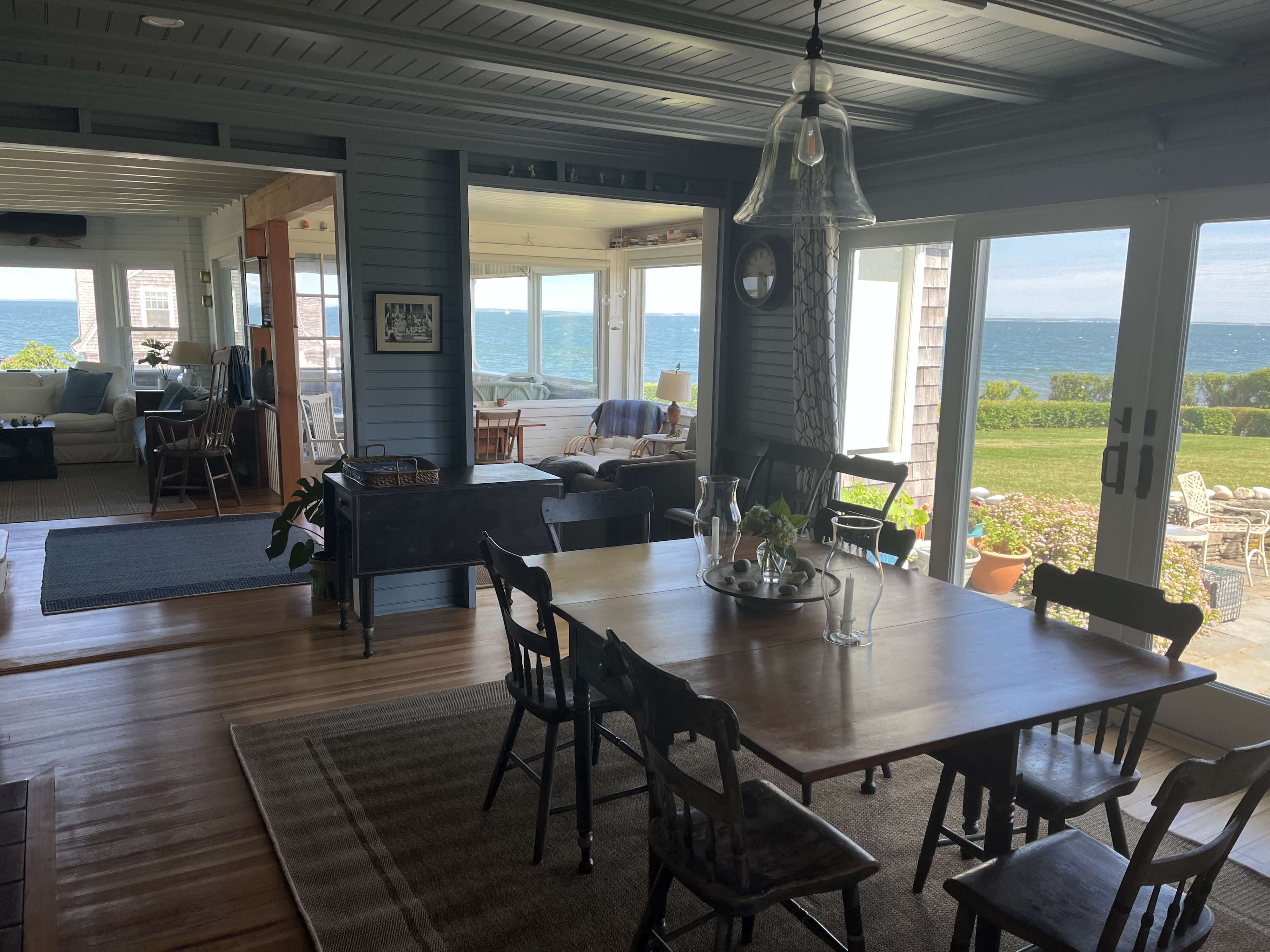 A bright dining area with a wooden table surrounded by chairs, overlooking a scenic view of the ocean through large glass doors.