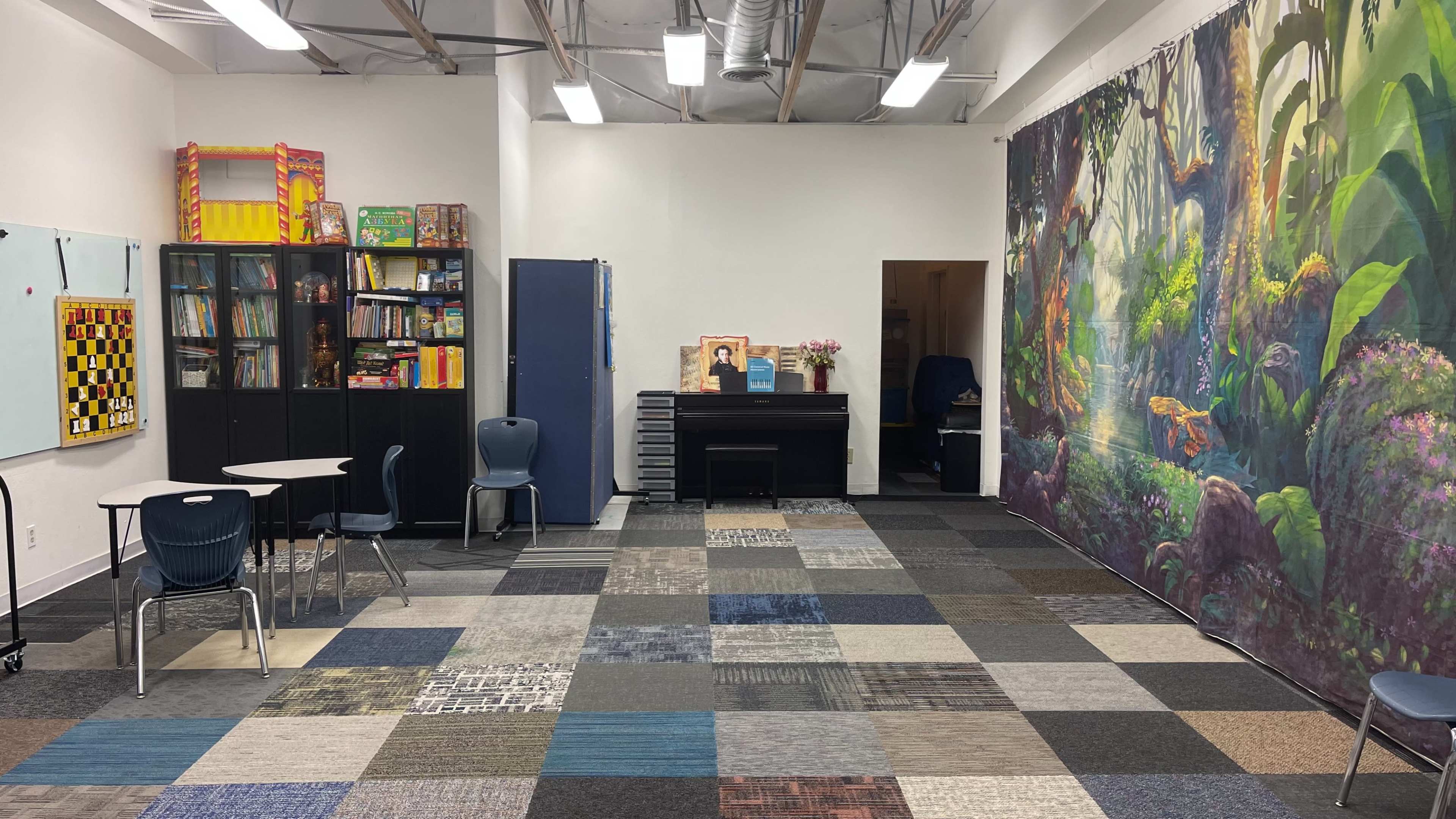 A classroom with a large mural of a forest, a central area of carpet tiles, a table and chairs, shelving filled with books, and a piano against one wall.