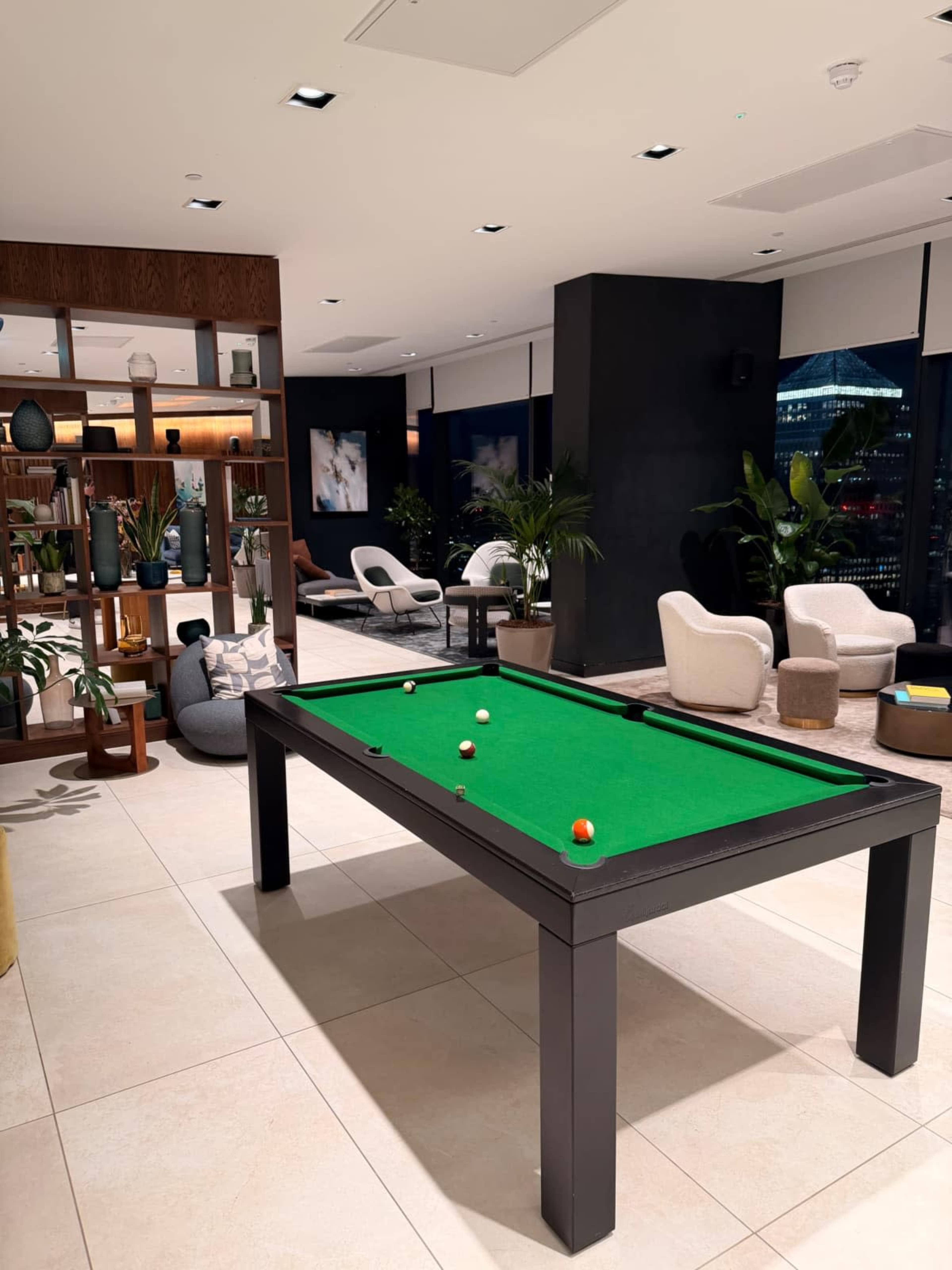 The image shows a modern lounge area featuring a green billiard table surrounded by seating and indoor plants.