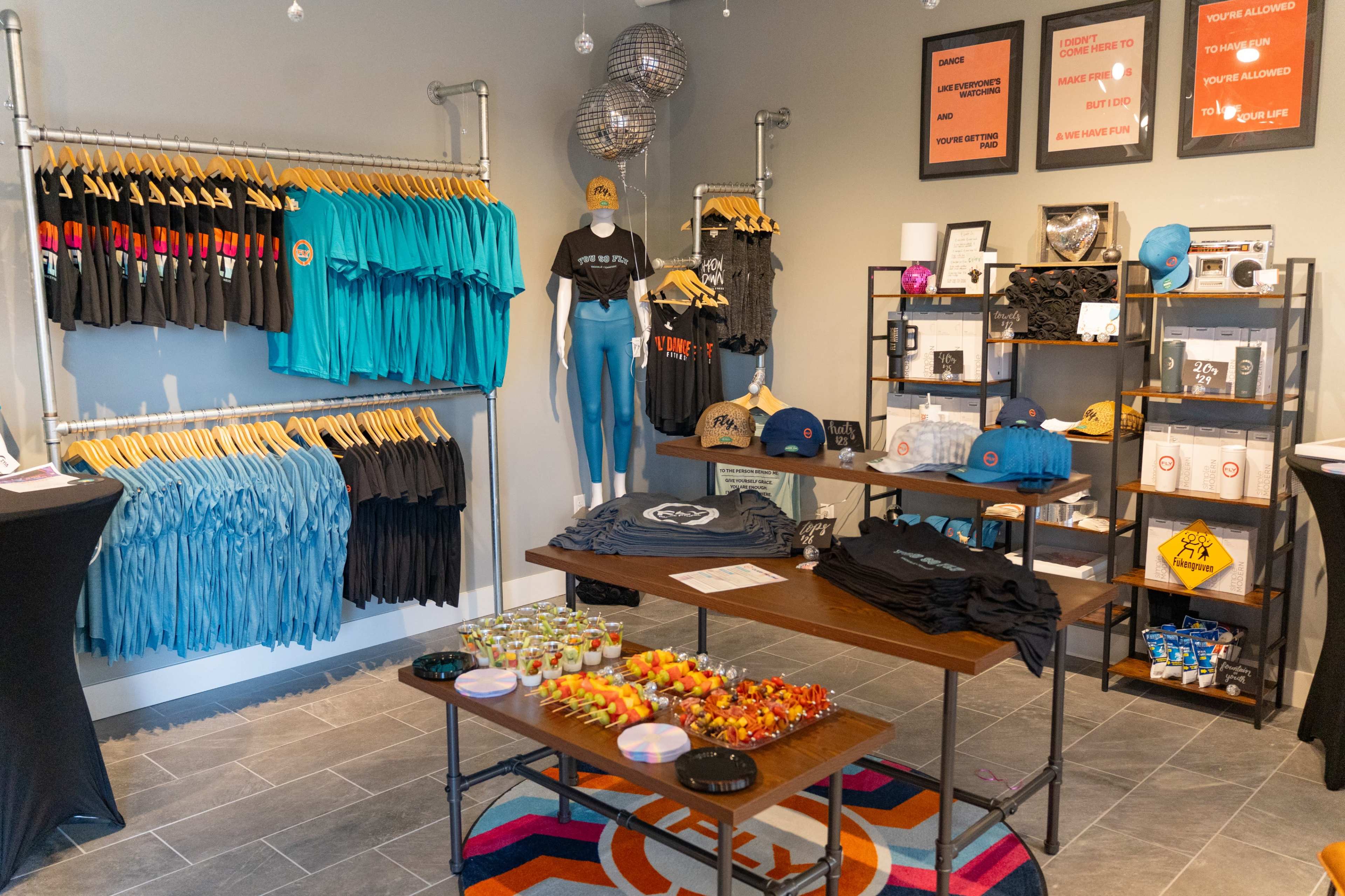 The image displays a retail space featuring various clothing items hanging on racks, a table with colorful snacks, and promotional materials on the walls.