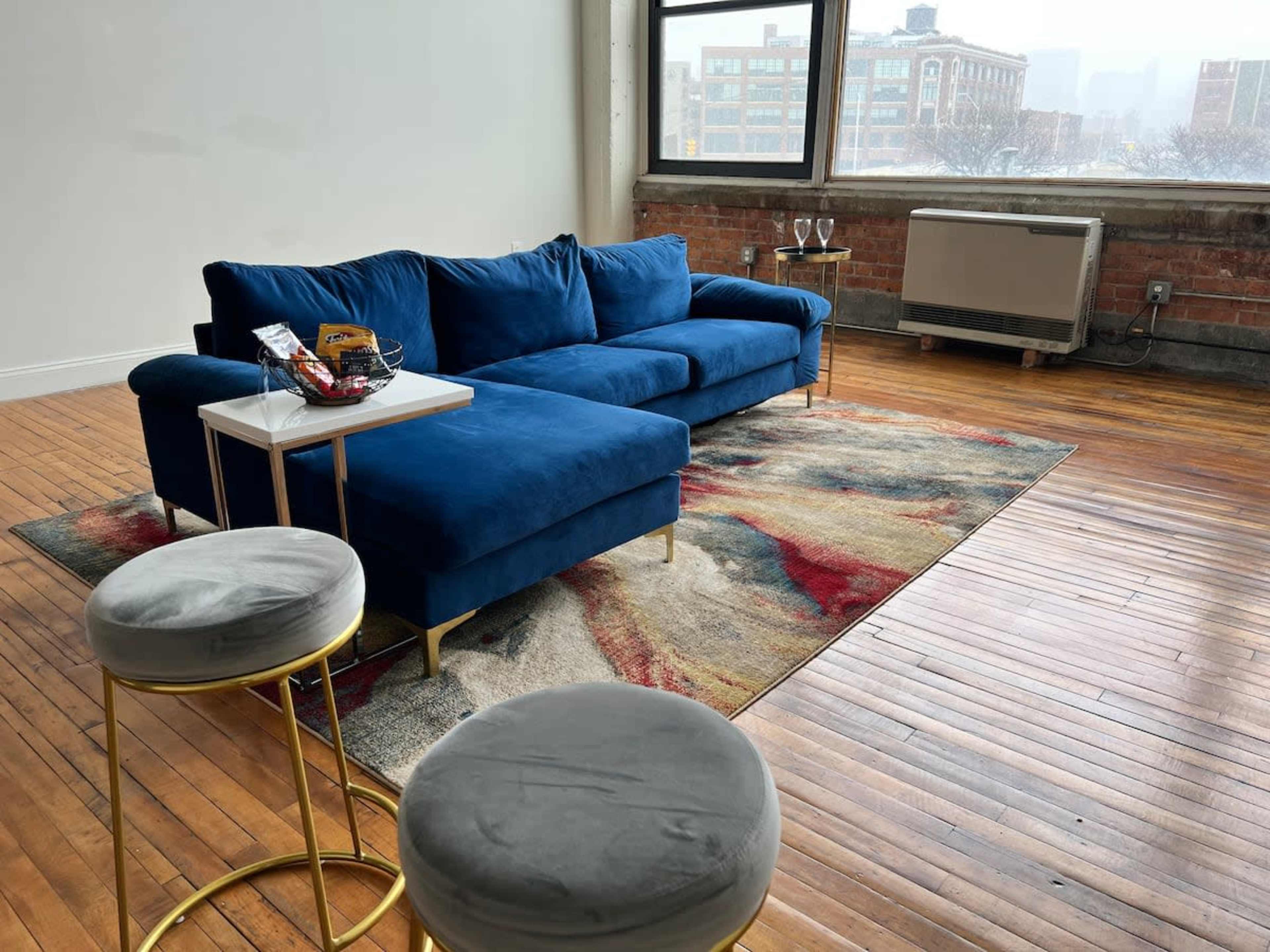 The image shows a blue sectional sofa on a colorful area rug in a room with large windows overlooking a cityscape, accompanied by two round stools and a small side table with snacks.