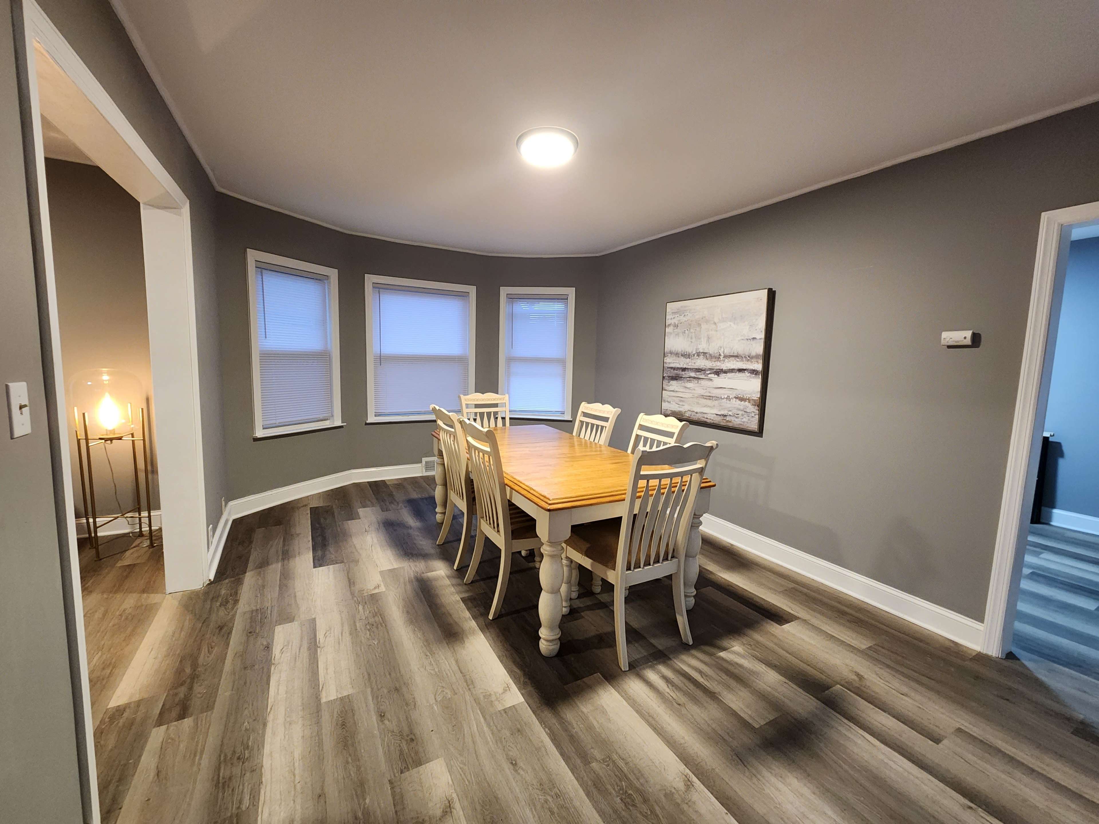 A dining room features a wooden table surrounded by six white chairs, with large windows and gray walls.