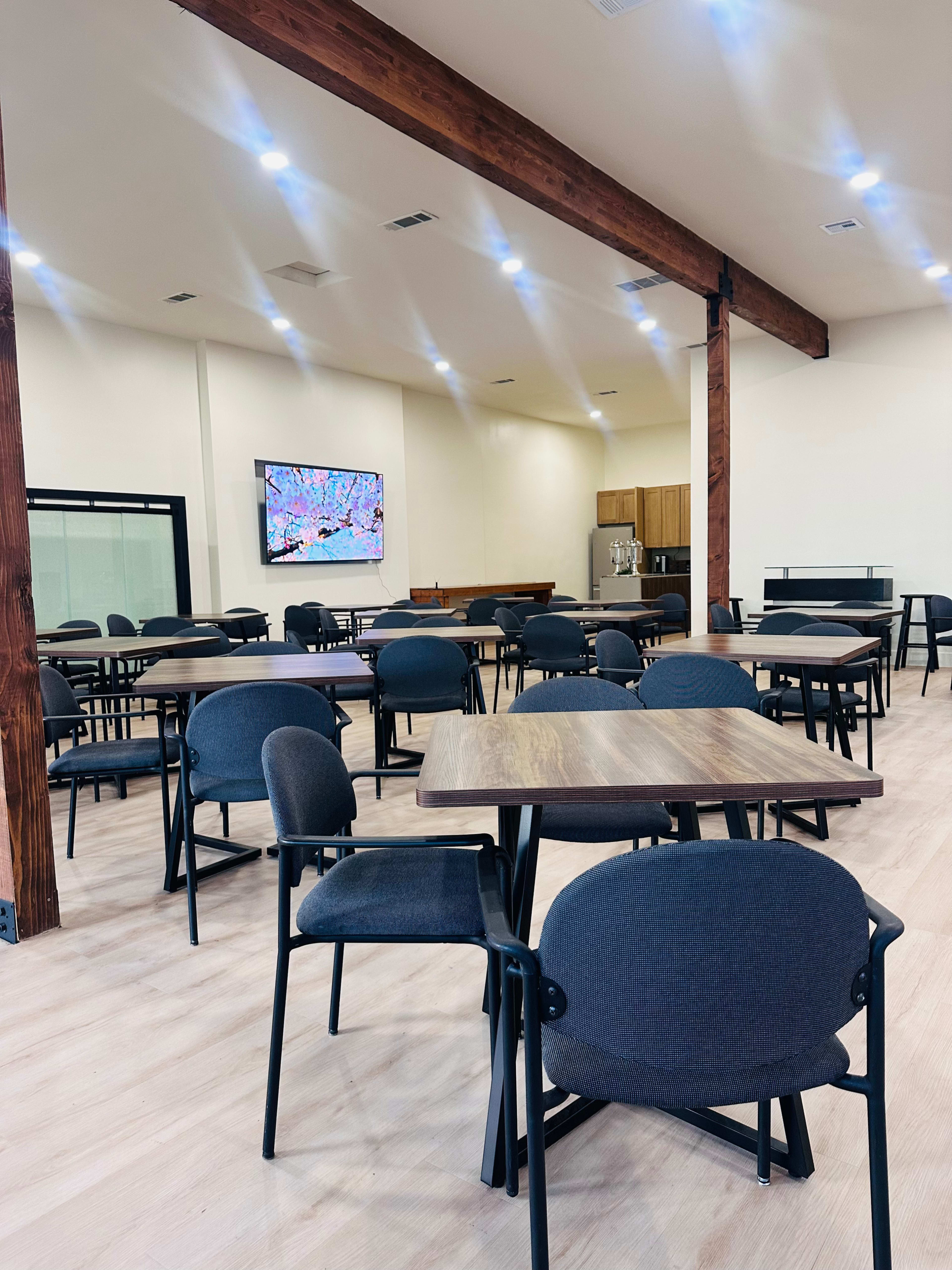 The image shows a spacious dining area with several wooden tables and chairs, decorated with wooden beams and equipped with a television screen on one wall.