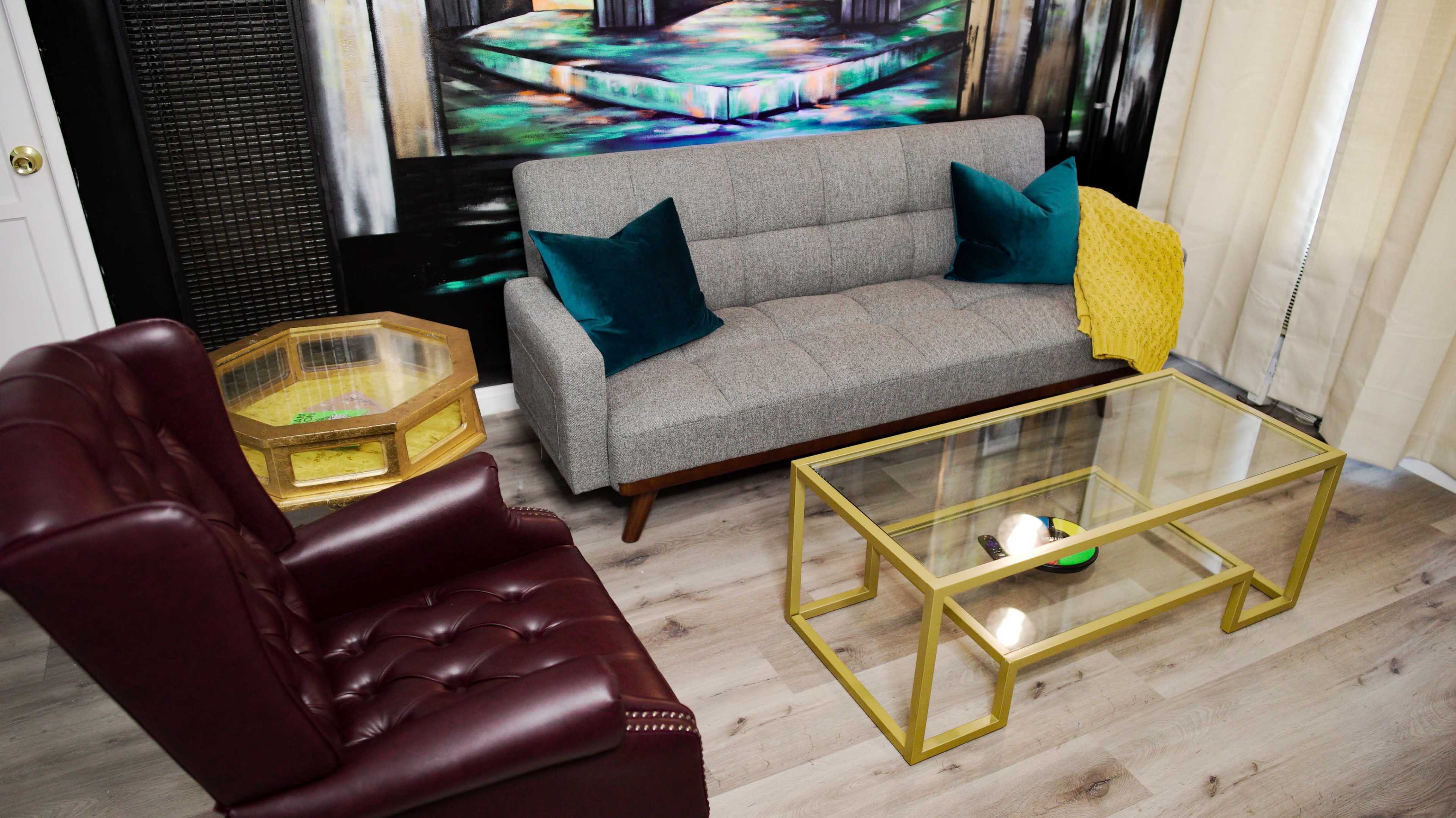 The image shows a modern living room setting with a gray sofa, two teal pillows, a maroon armchair, and a glass coffee table on light wooden flooring.