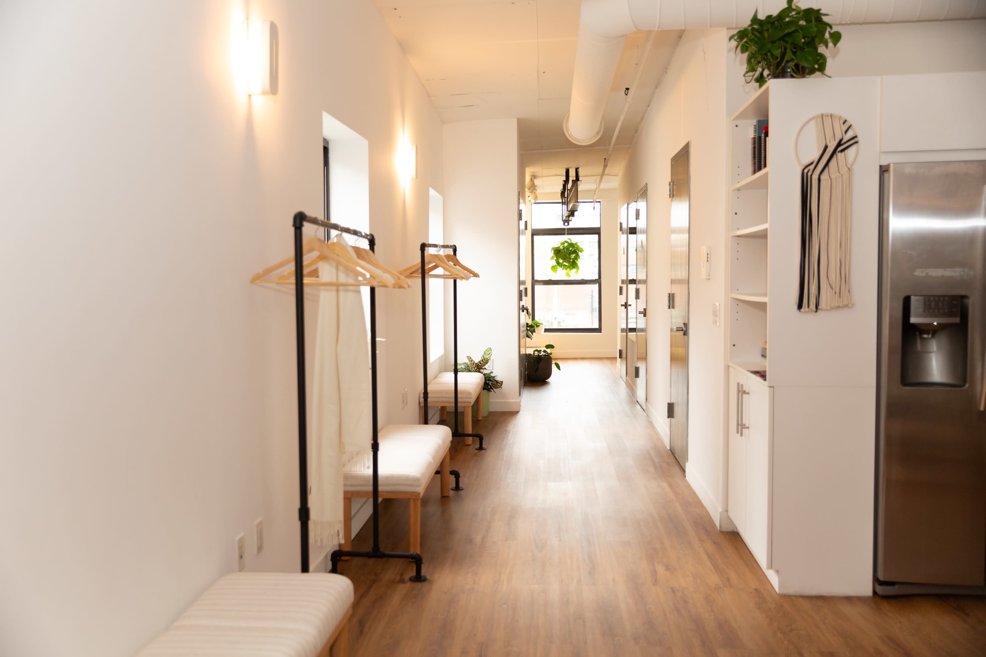 The image shows a well-lit hallway with minimalist decor, featuring wooden flooring, two benches, clothing racks, and plants along the walls.