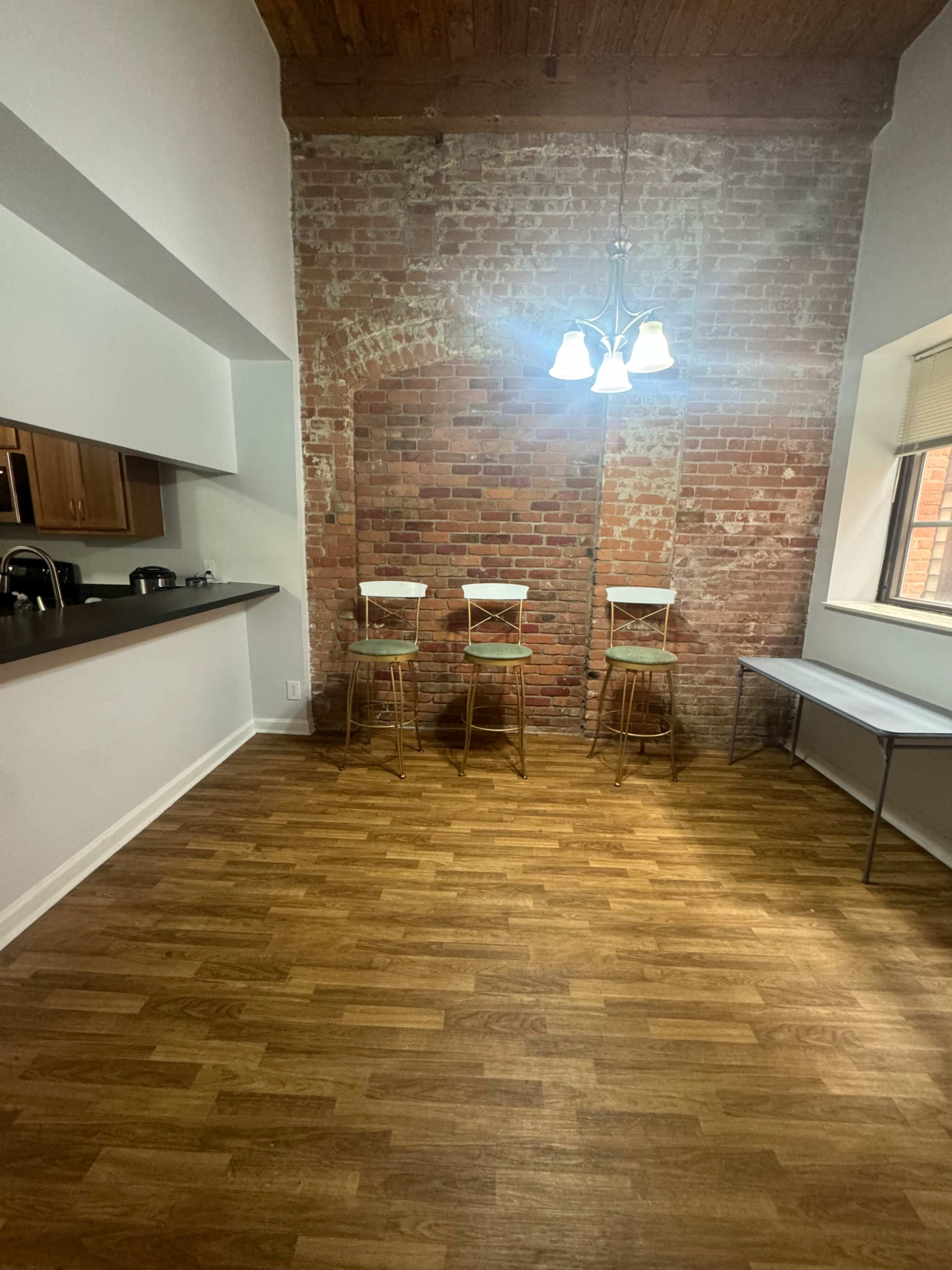 The image shows an empty room featuring a brick wall, four wooden bar stools, a small table, and a light fixture.