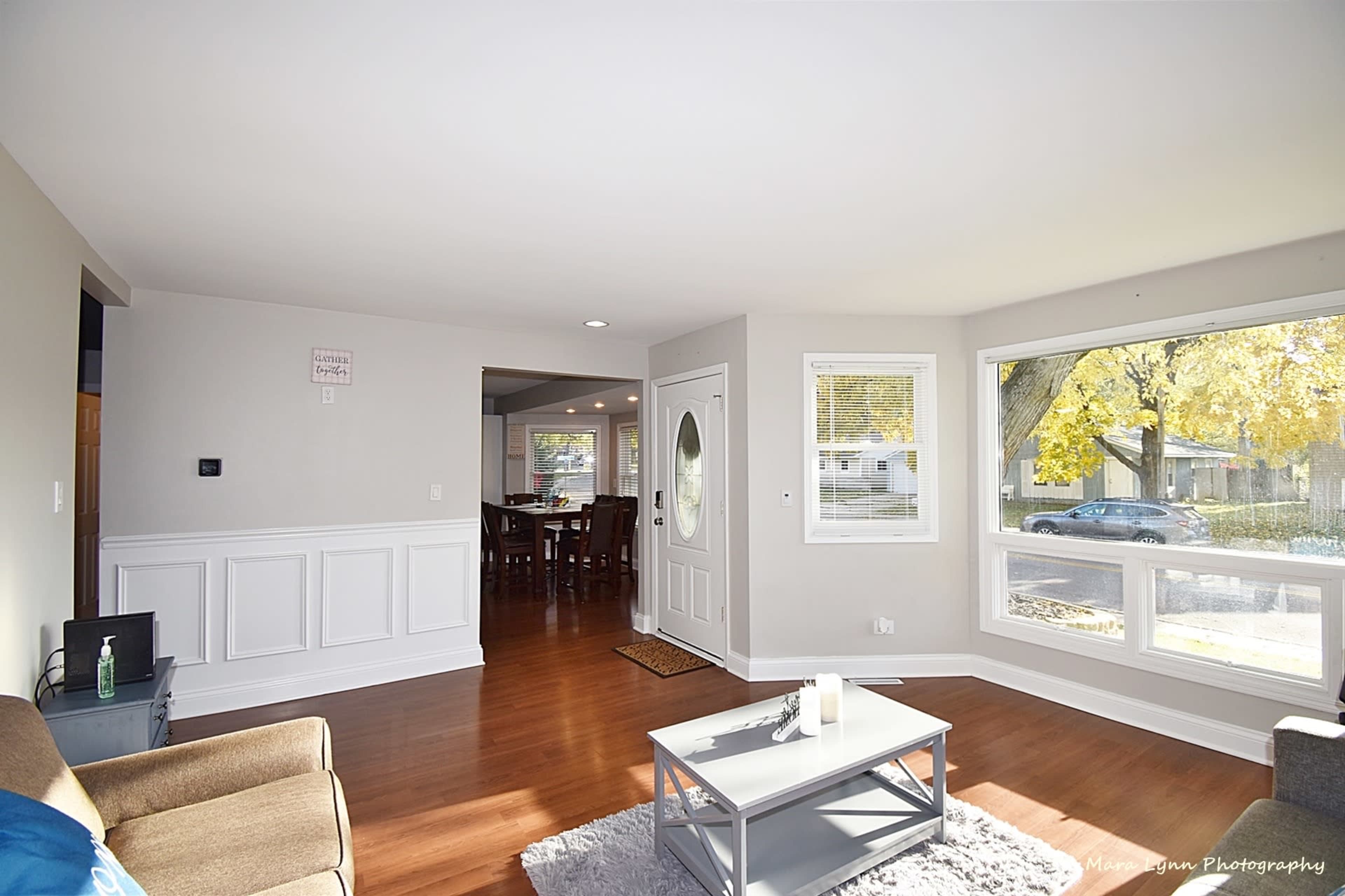 The image shows a bright living room with hardwood floors, a sofa, and large windows overlooking a yard.