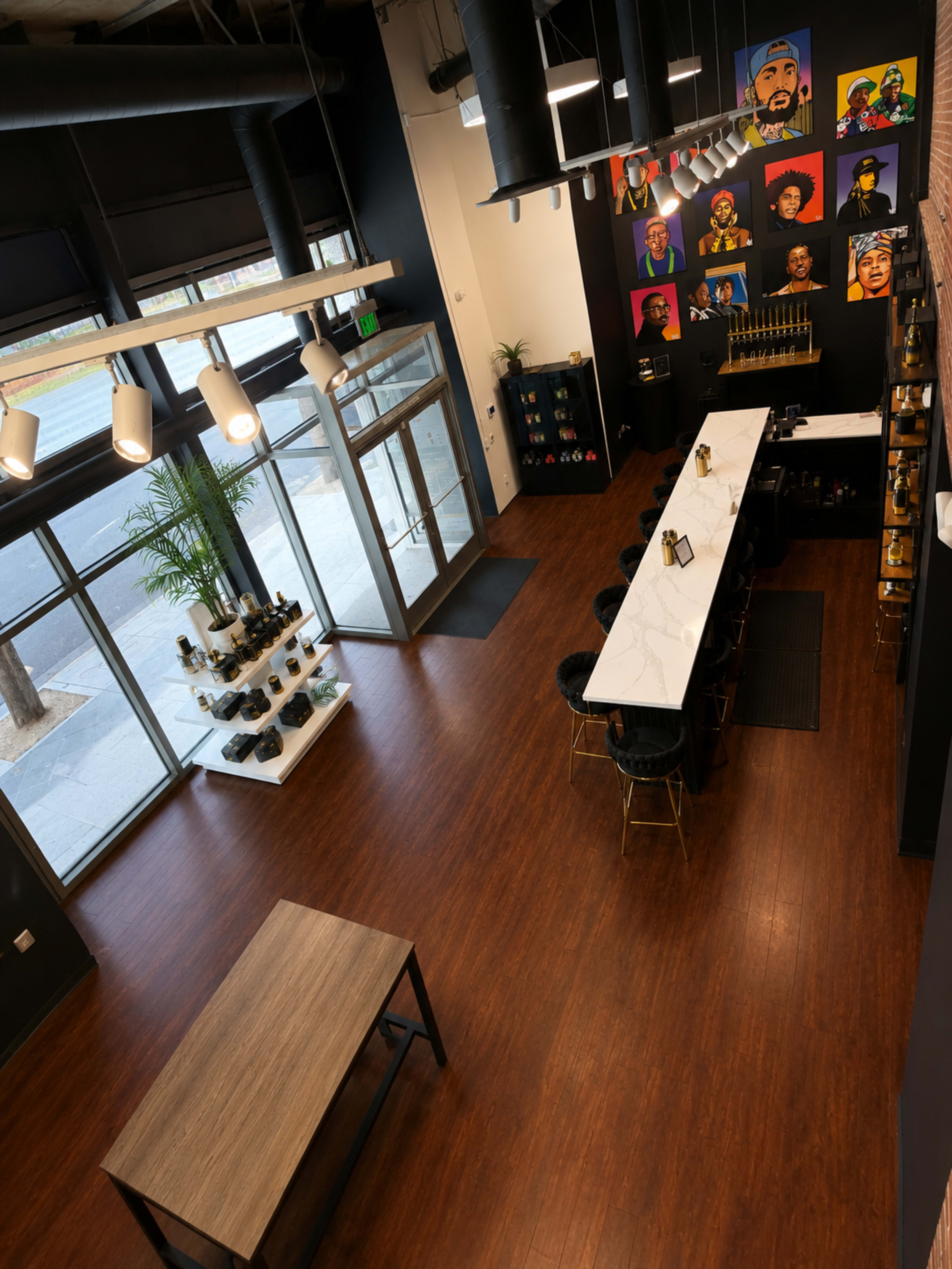 The image shows a modern retail space with a bar-like counter, a wooden table, artwork on the walls, and large glass entrance doors allowing natural light to enter.