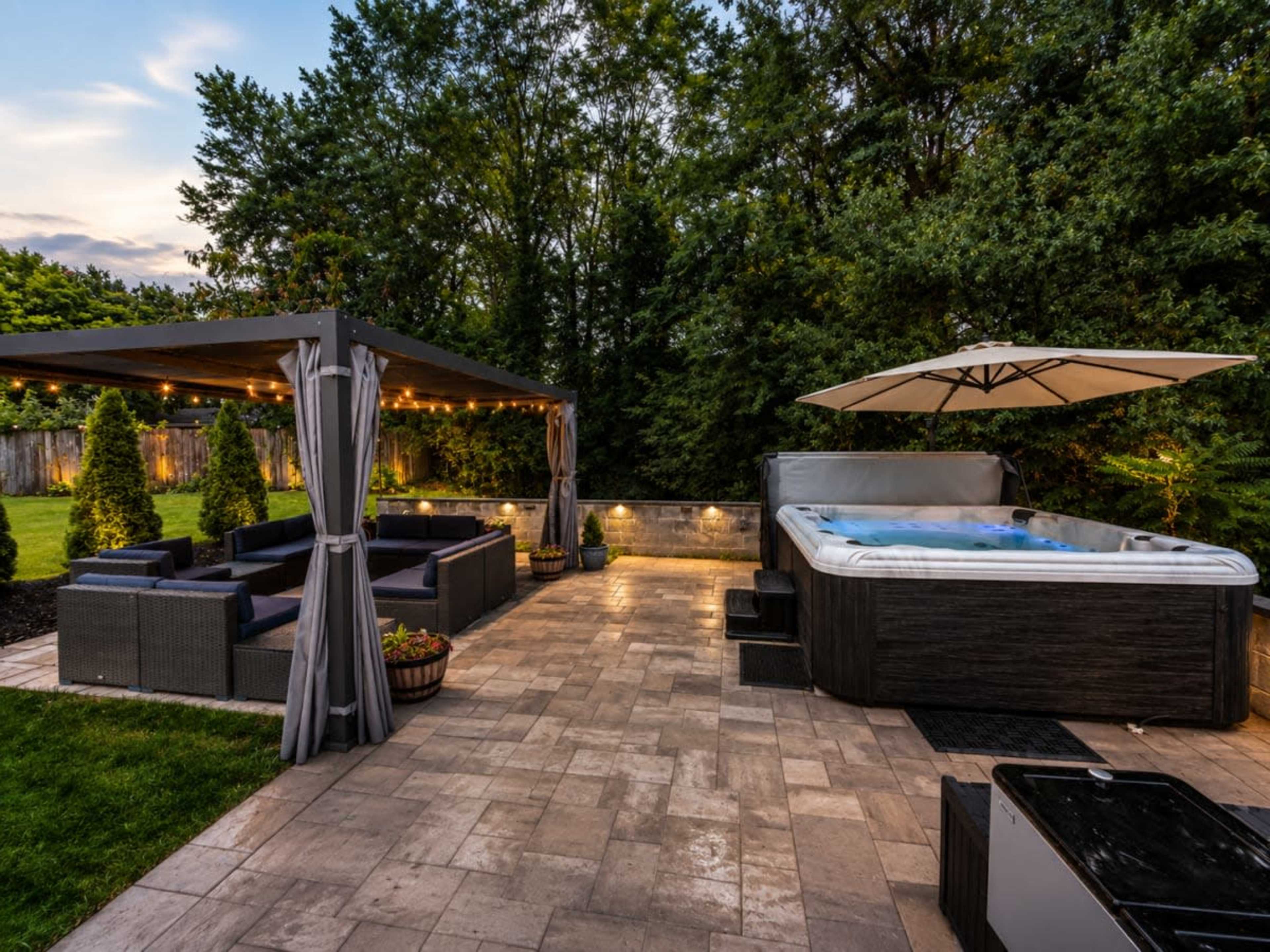The image shows a backyard patio featuring a hot tub, a seating area with a gazebo, and landscaped greenery.