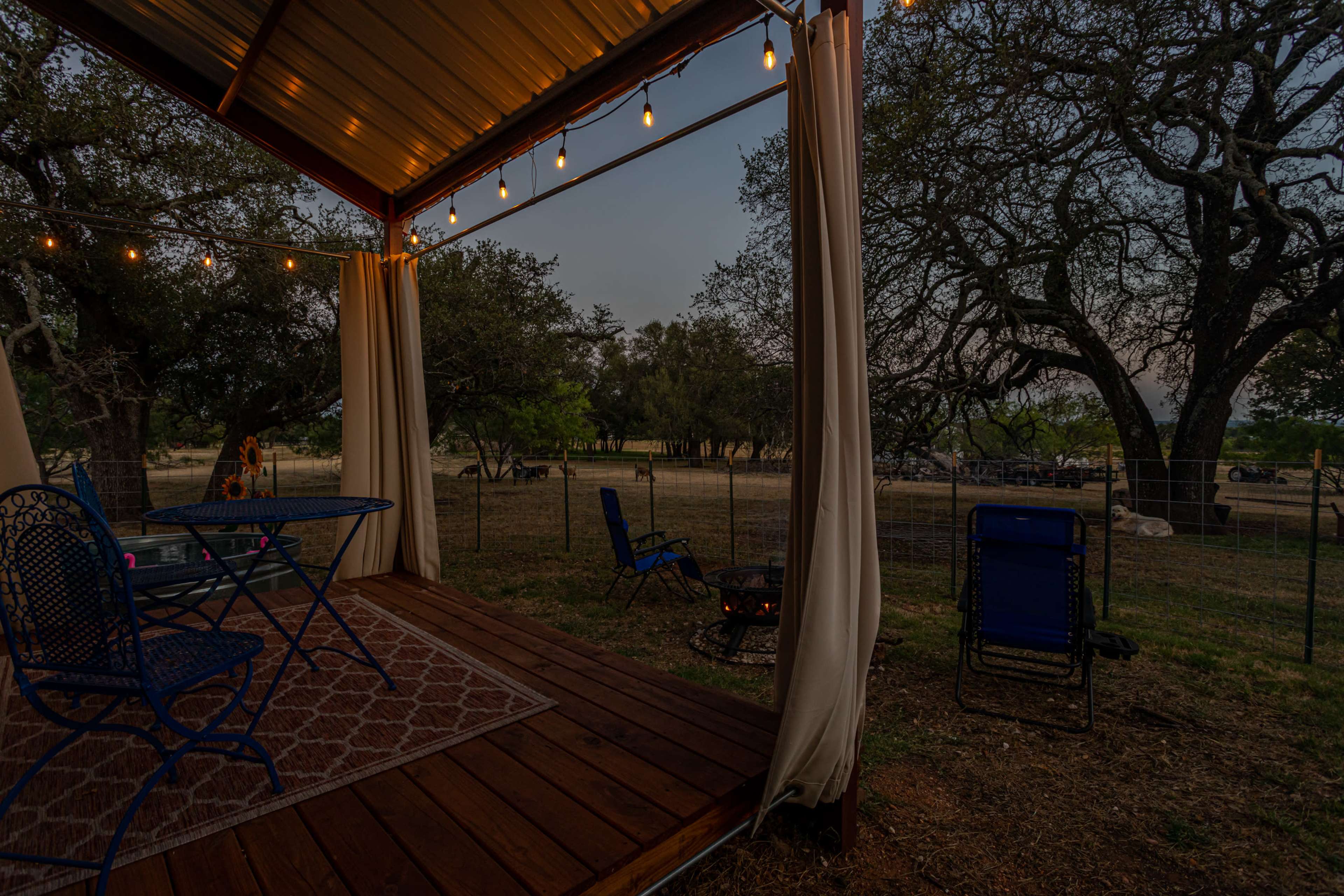 A wooden deck with string lights and a table and chairs overlooks a grassy area with trees and a fence in the distance.