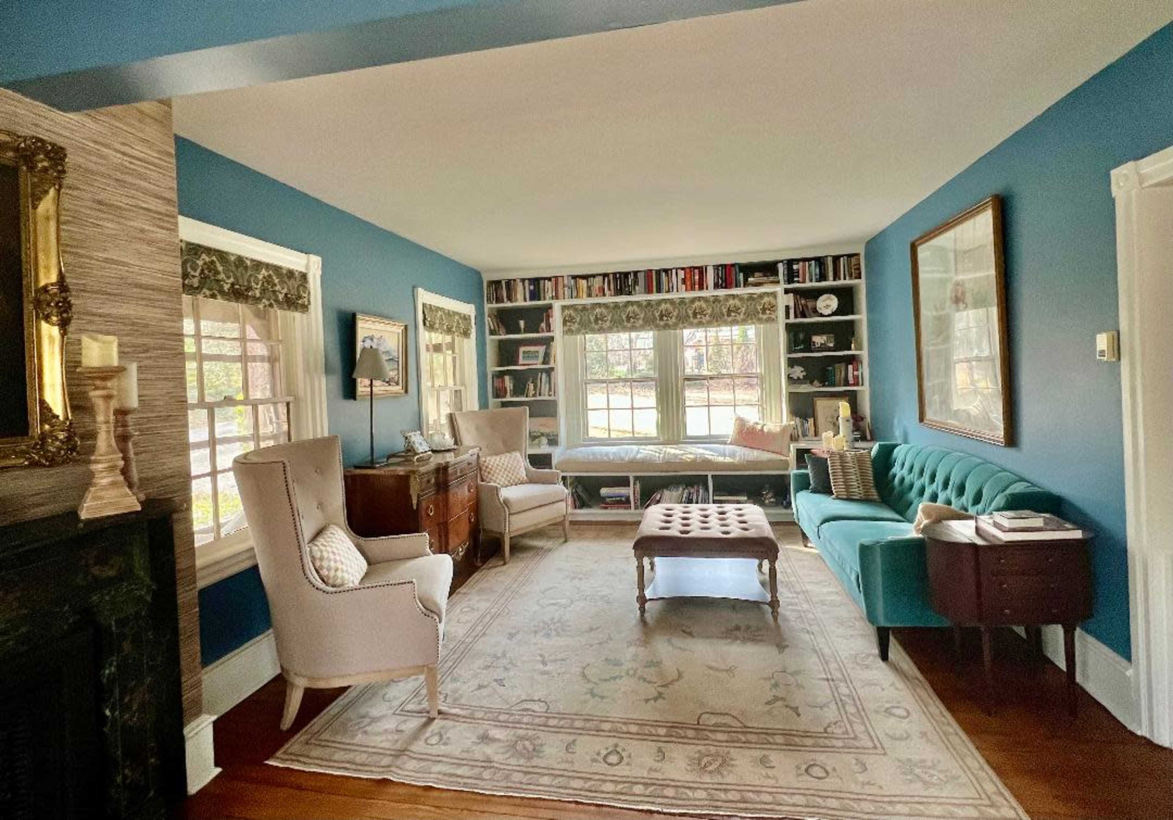 The image shows a cozy living room with blue walls, a patterned rug, a teal sofa, two upholstered chairs, and a large window lined with bookshelves.