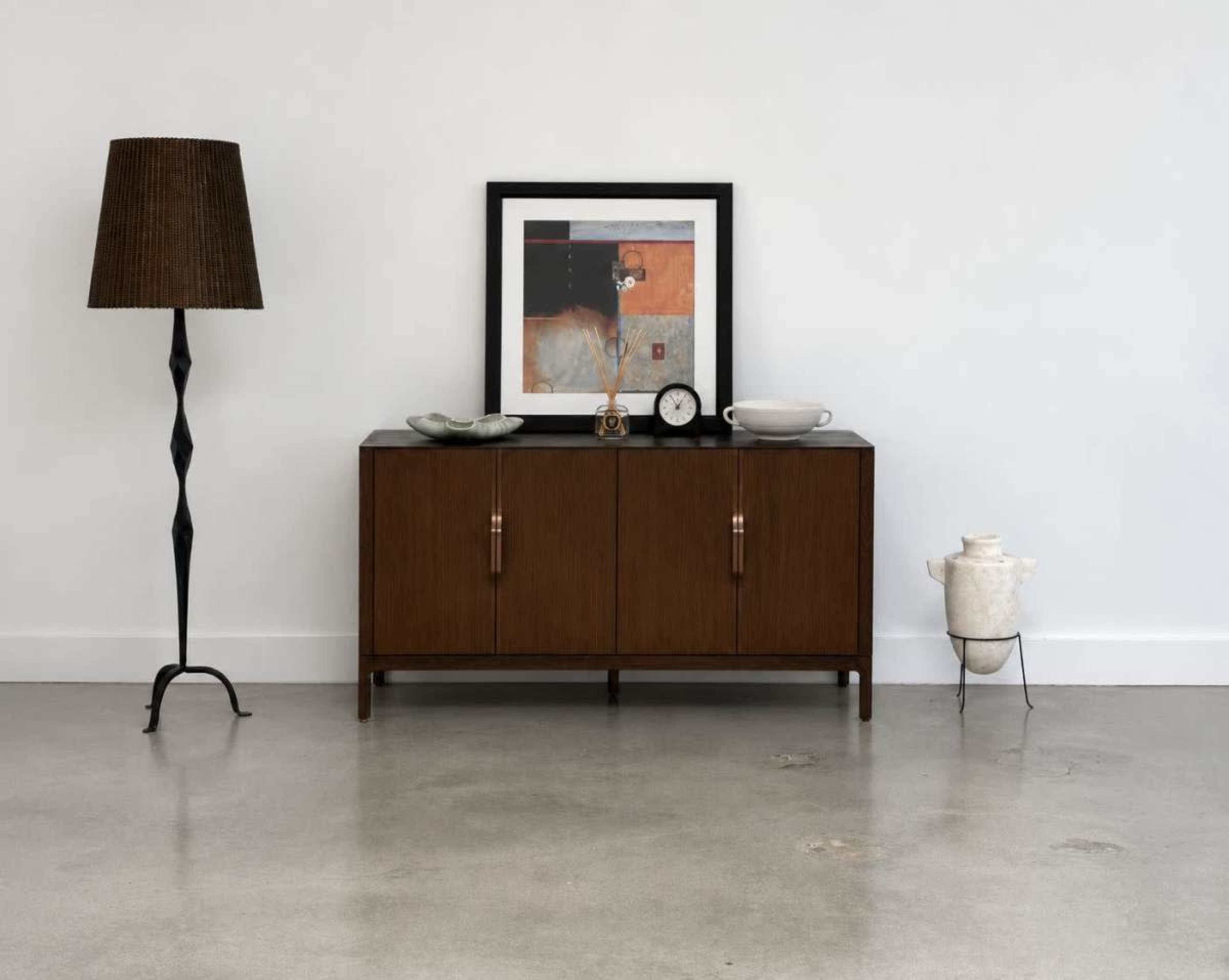 The image shows a mid-century modern wooden sideboard with two doors, a decorative clock, a bowl, and a framed abstract artwork above, accompanied by a woven floor lamp and a white ceramic teapot on the floor.