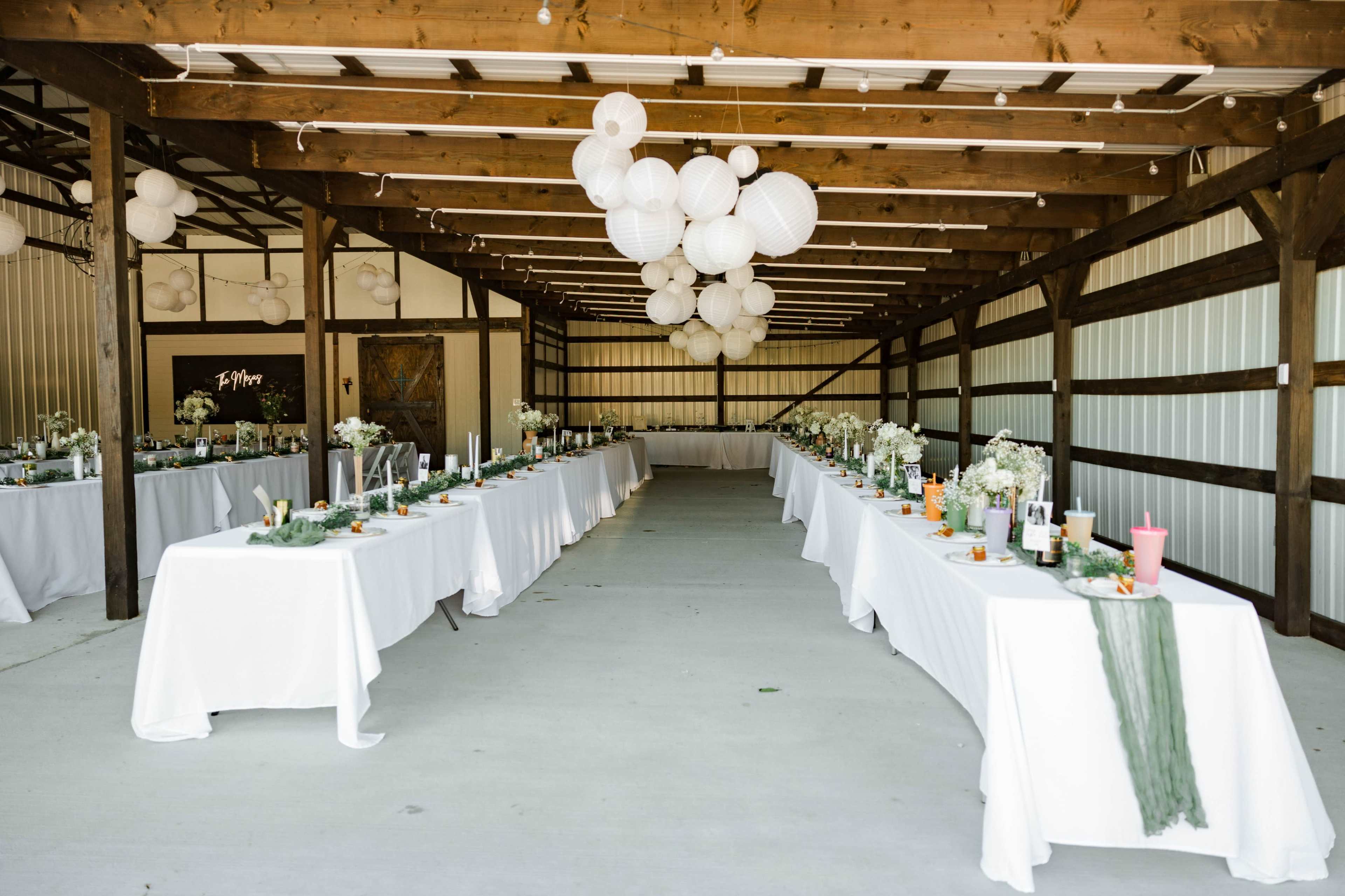 A spacious barn setting features two long tables adorned with white tablecloths and floral centerpieces, illuminated by decorative paper lanterns overhead.