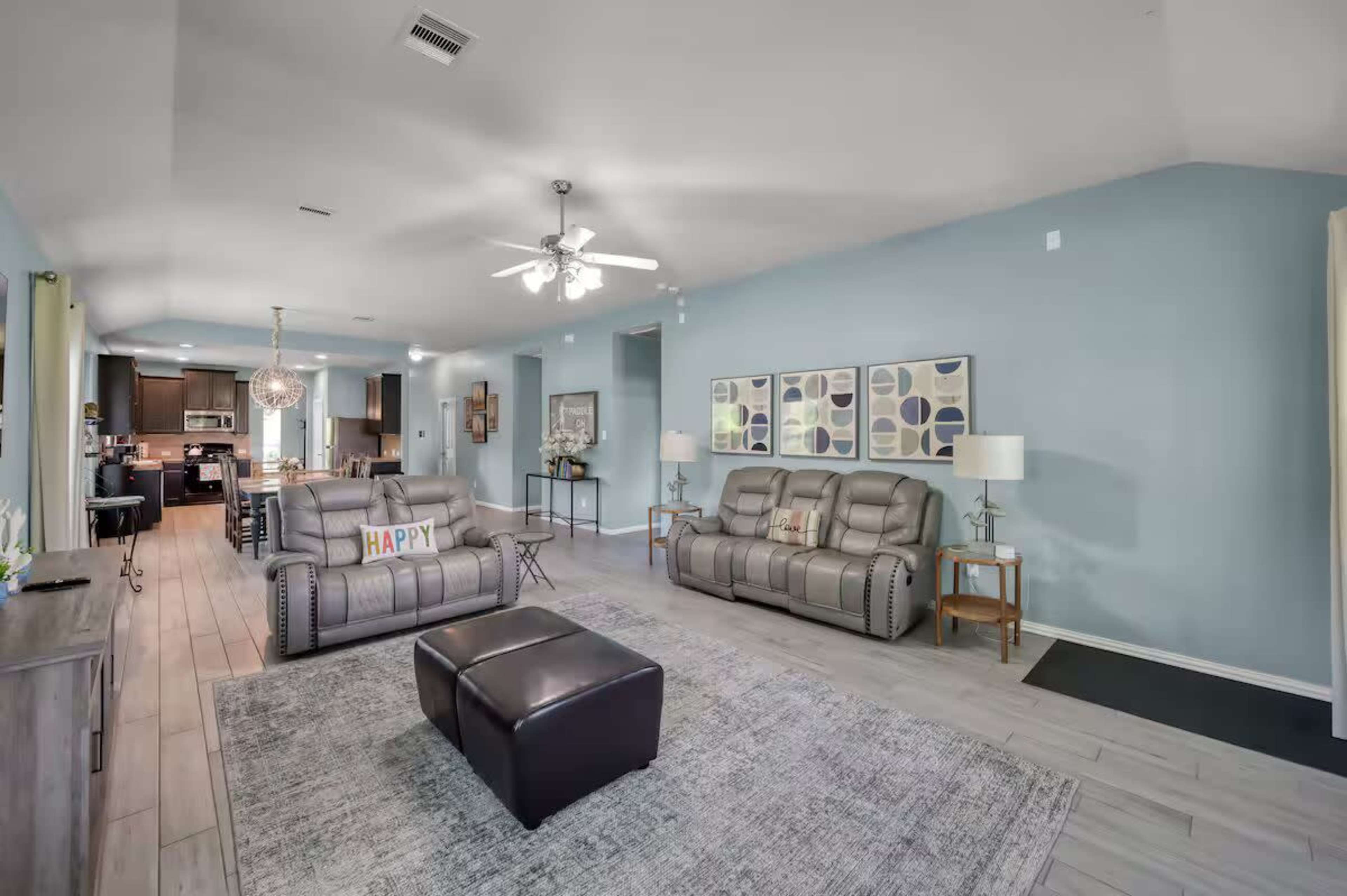 The image shows a spacious living room with two leather couches, a coffee table, and a light blue wall, leading into a kitchen area in the background.