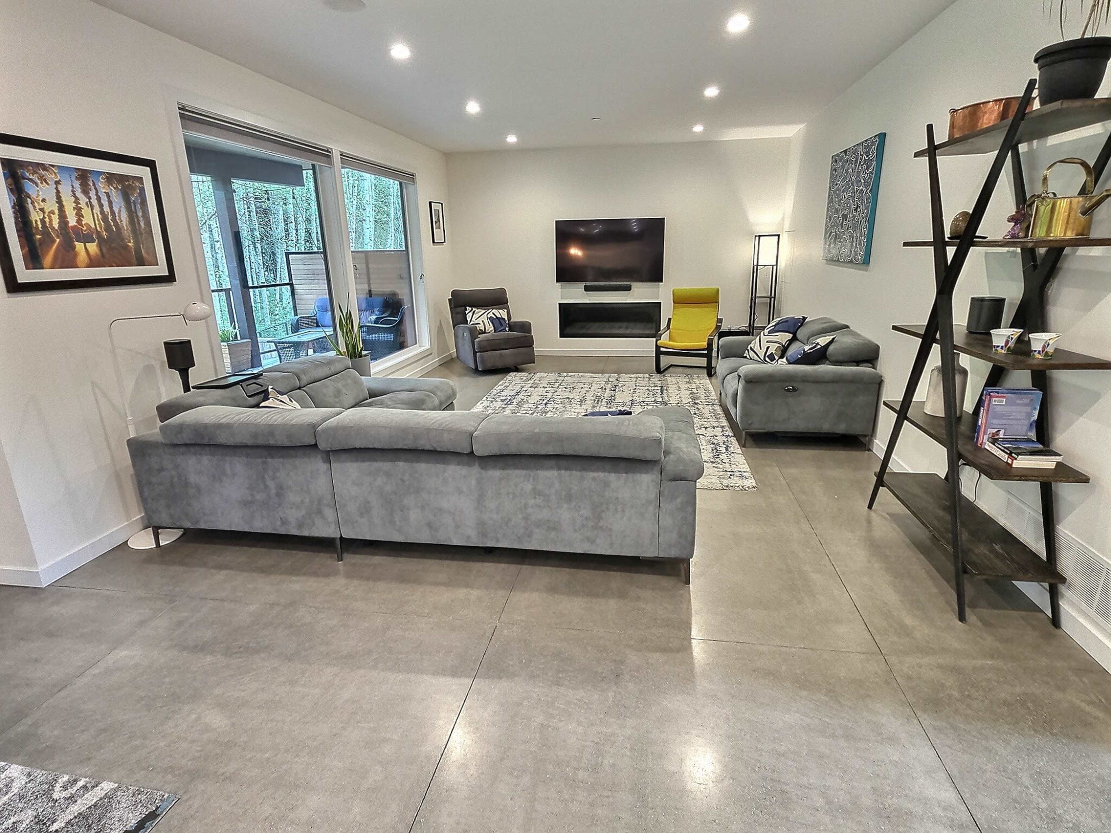 A modern living room with gray furniture, a television mounted on the wall, and large windows facing an outdoor area.