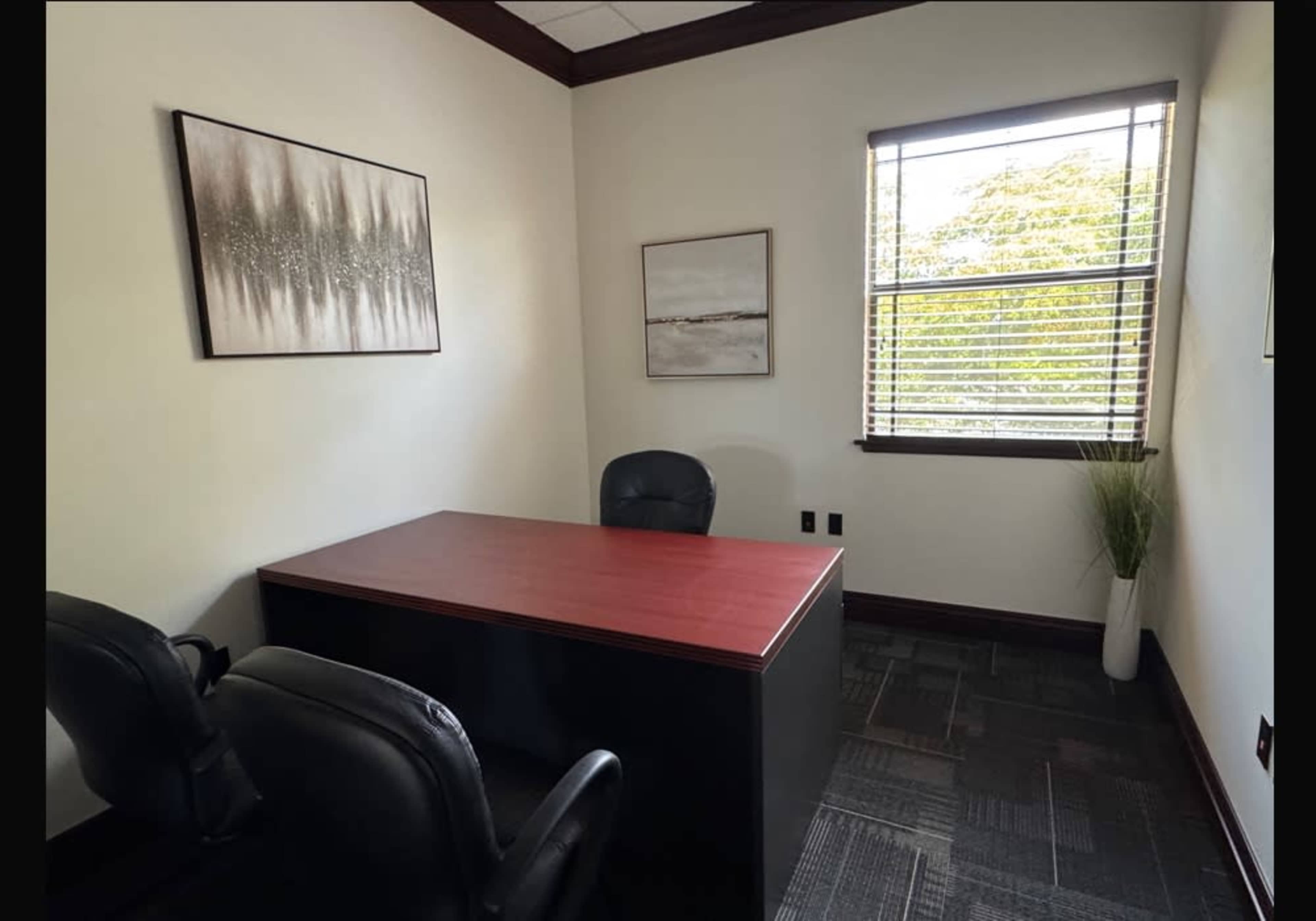 The image shows a small office with a wooden desk, two black chairs, and two framed artworks on the walls.
