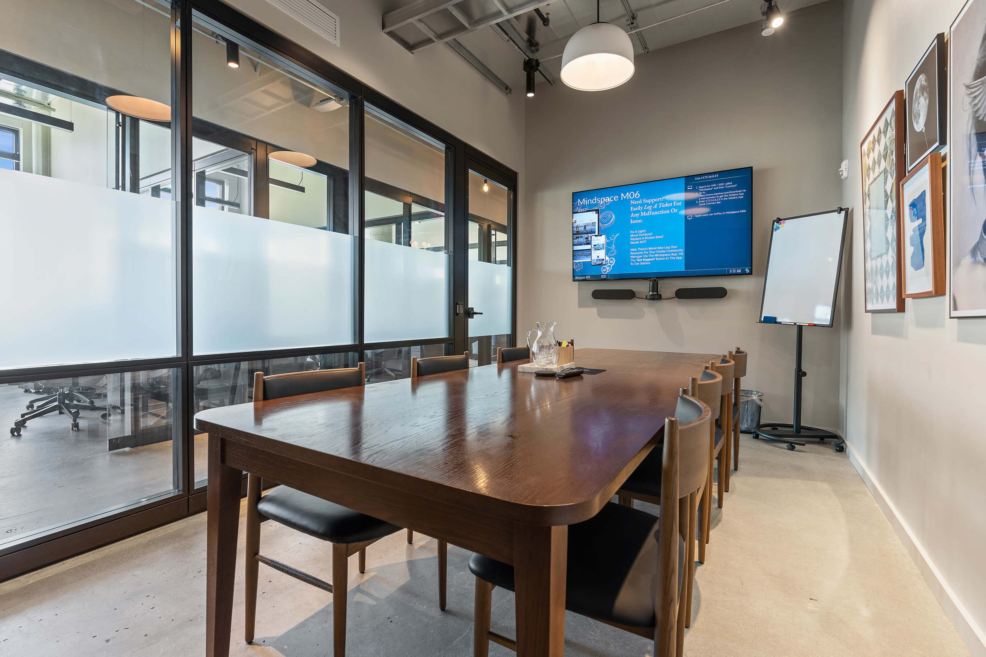 A modern conference room features a long wooden table, chairs, a wall-mounted screen, and glass walls allowing visibility into a neighboring workspace.