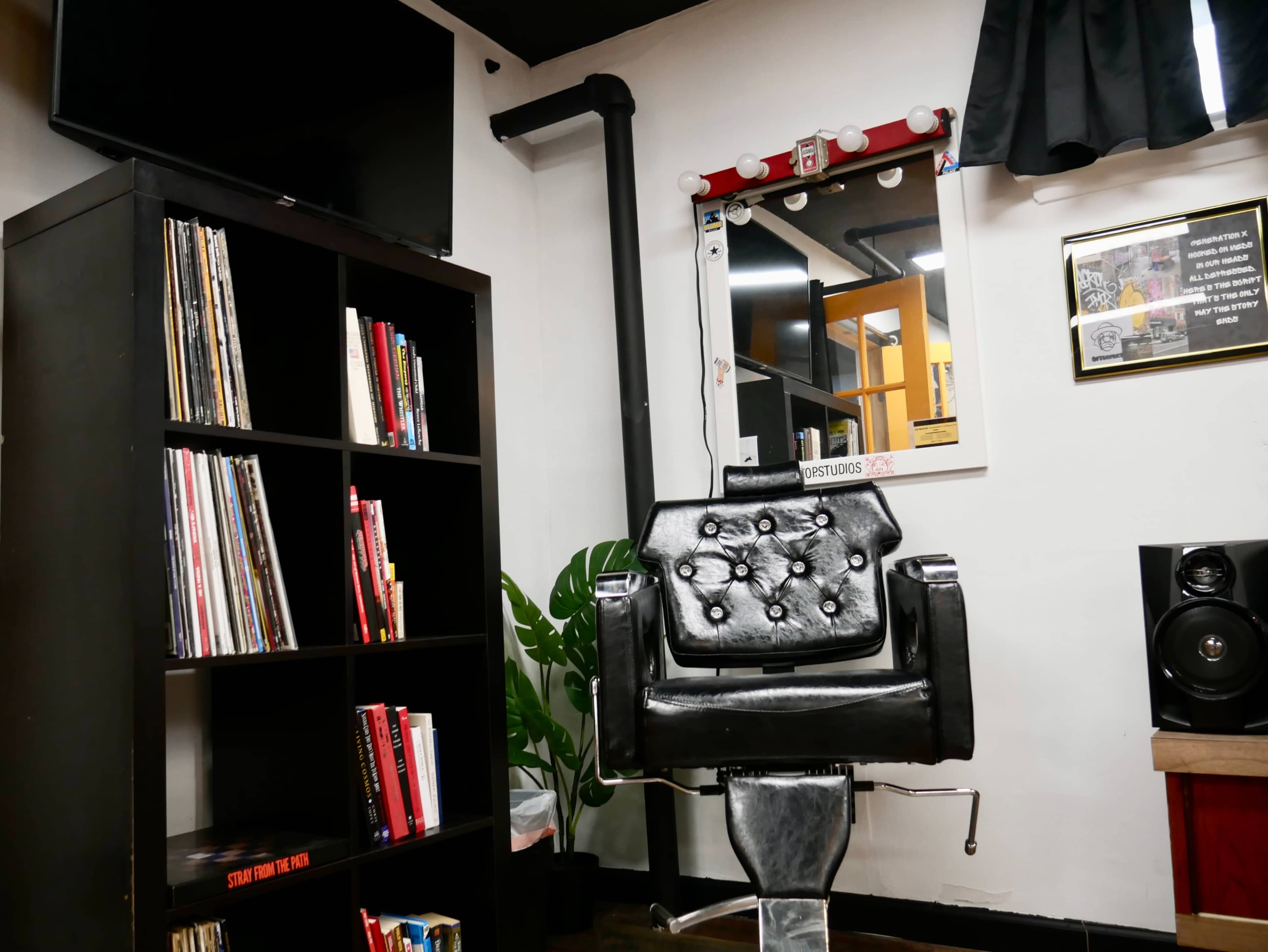 The image shows a black barber chair positioned in a well-lit barbershop, next to a bookshelf filled with books and vinyl records, and a mirror reflecting the shop's interior.