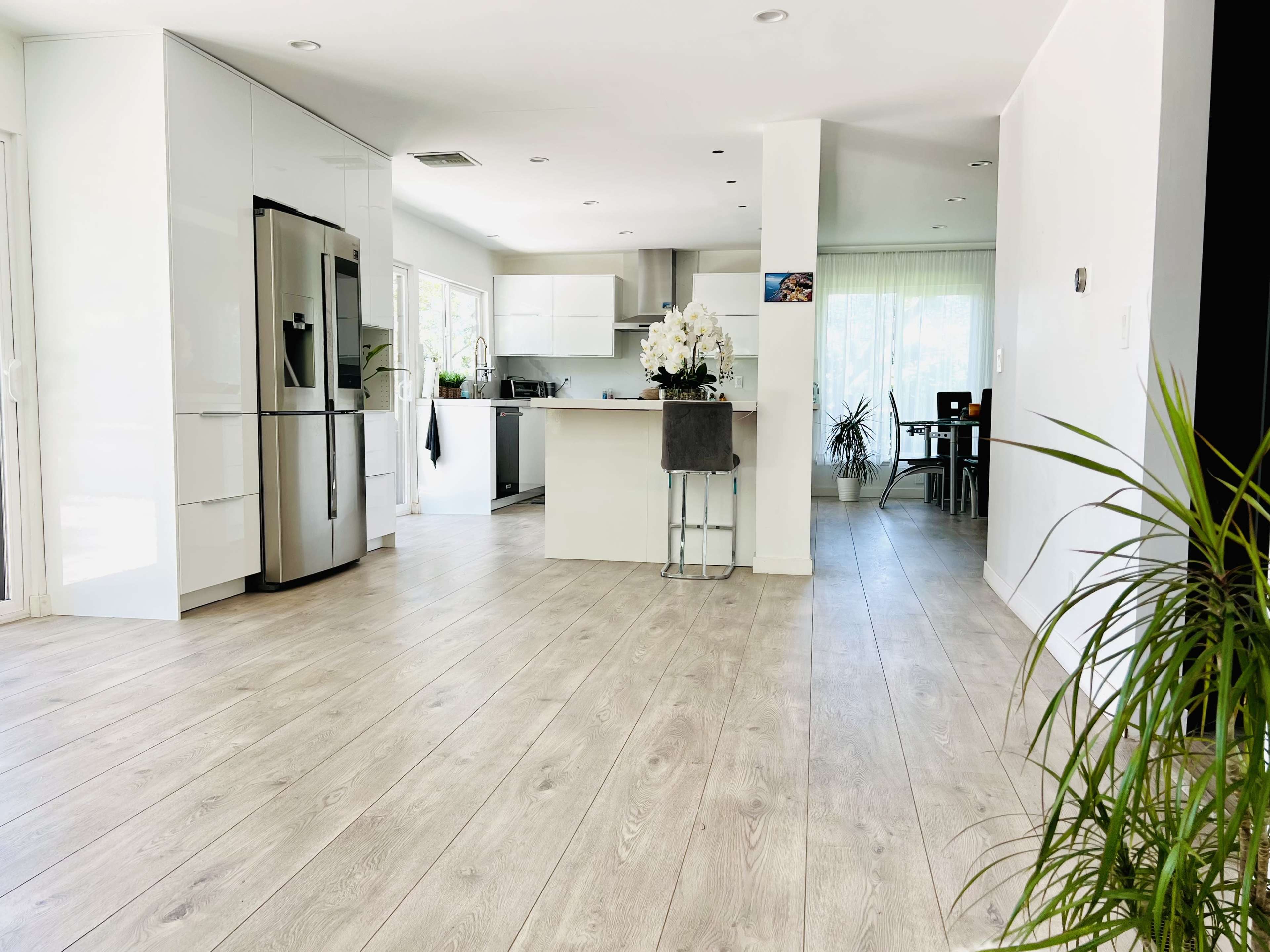 The image depicts a modern kitchen and dining area with light wood flooring and bright natural lighting.