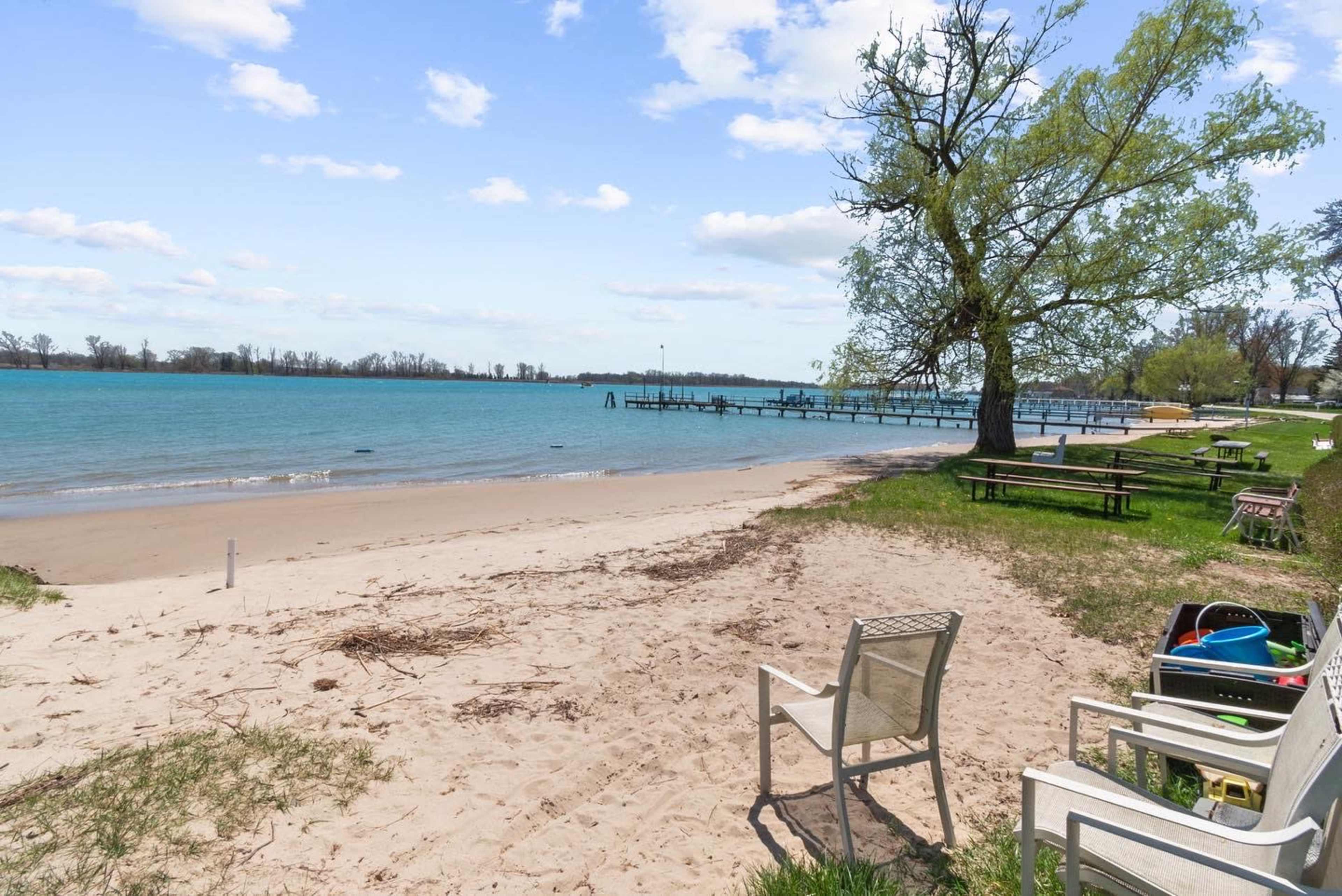 A sandy beach is bordered by a calm body of water, with a wooden pier extending into the lake and picnic tables set up on the grassy area nearby.