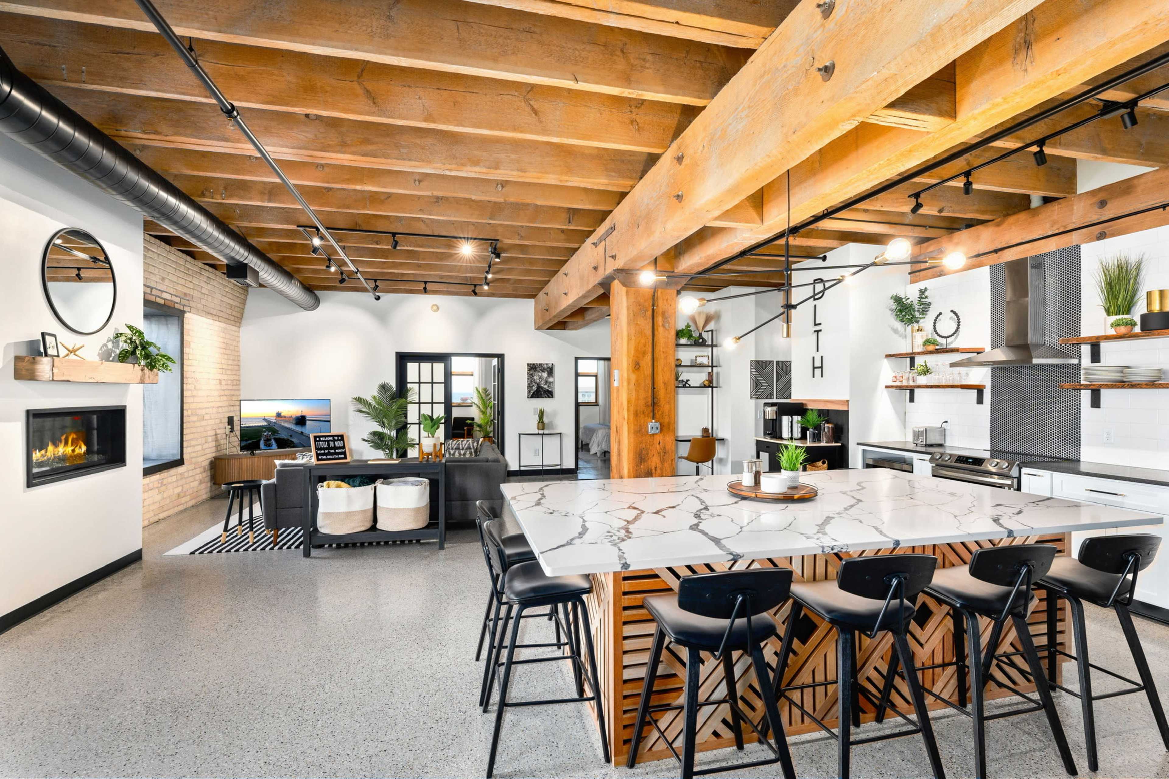 A modern kitchen with a large marble island, black bar stools, wooden beams, and open space leading into a living area with a fireplace and decorated walls.