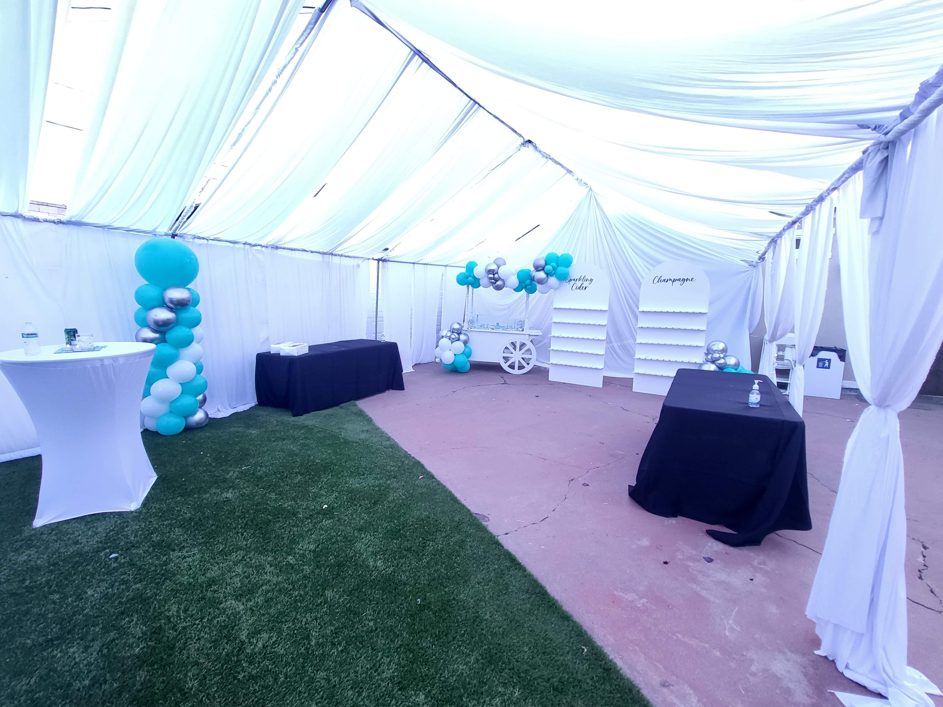 The interior of a tented event space features two black tables, one white table with a decorative balloon installation, and a grass-like floor covering.