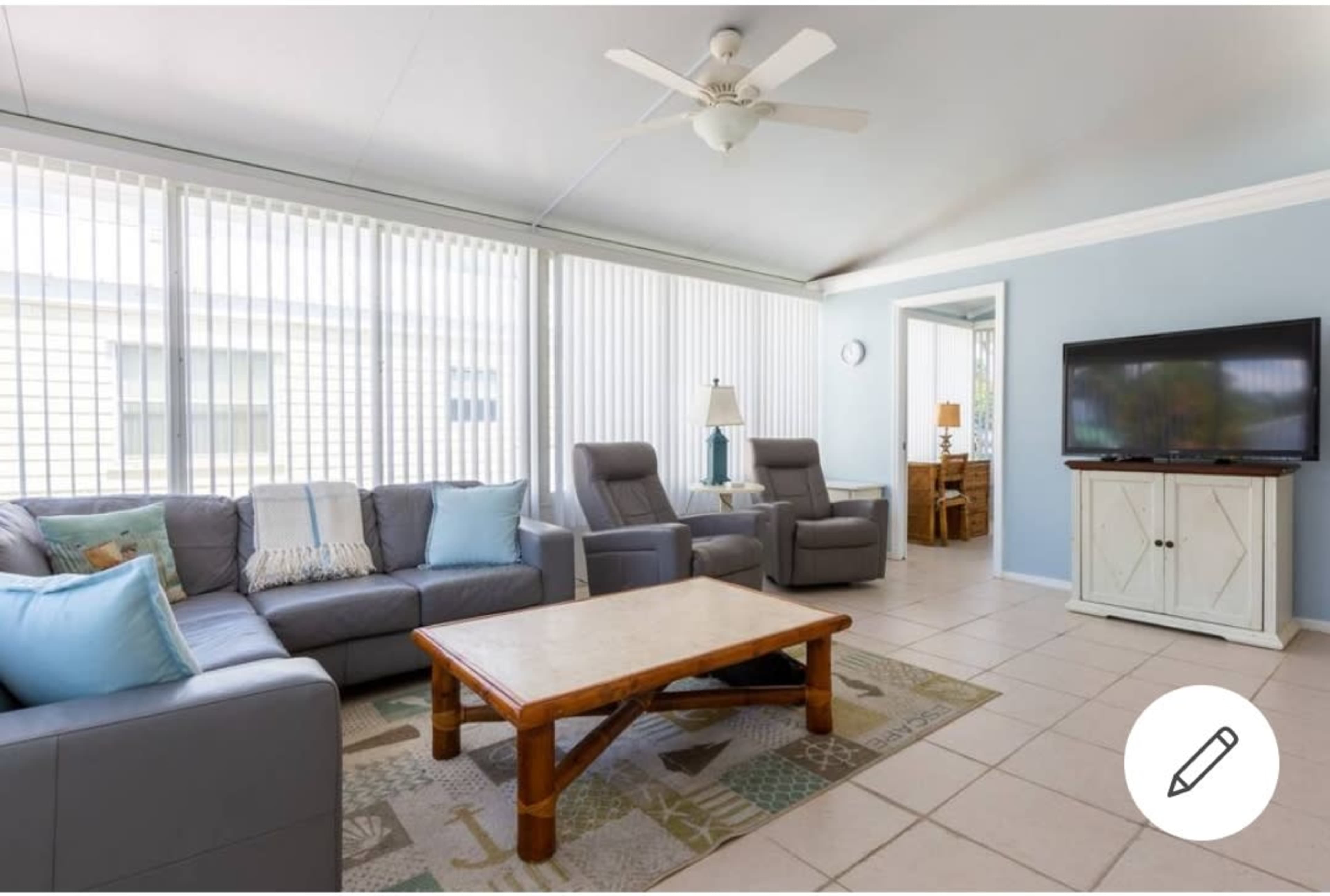 A bright living room with a gray sectional sofa, two recliners, a wooden coffee table, and a television against a light blue wall.