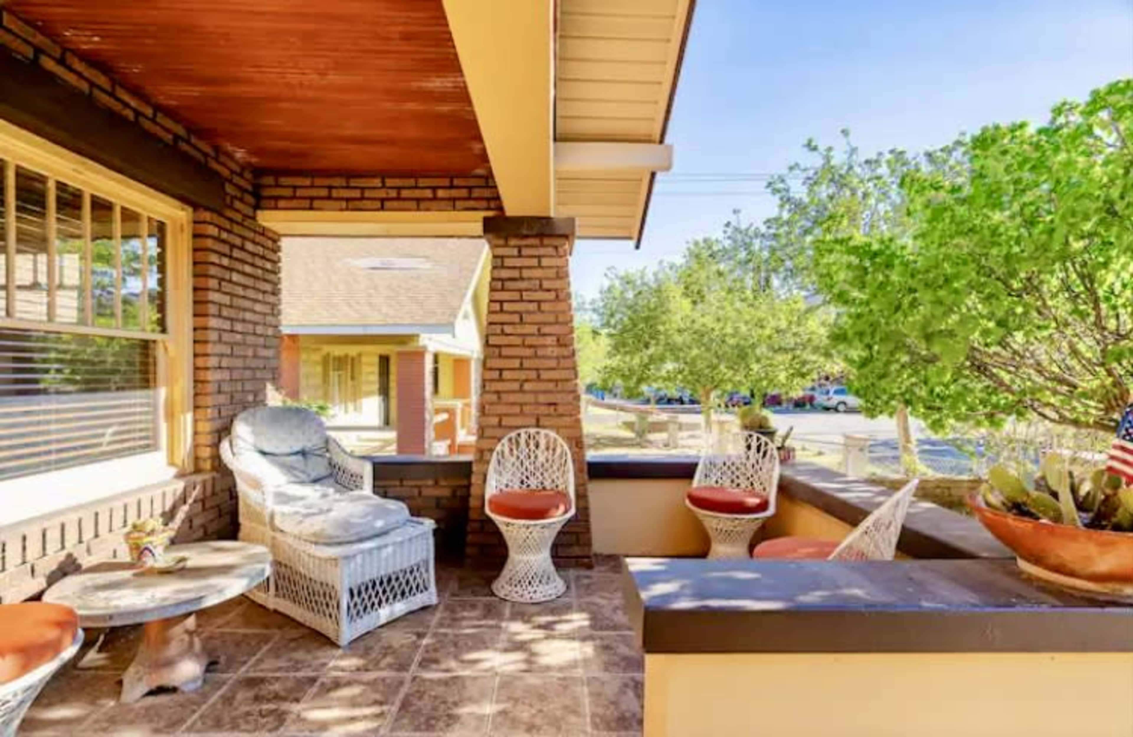 A porch with wicker furniture, a small table, and potted plants, surrounded by trees and a view of parked cars in the background.