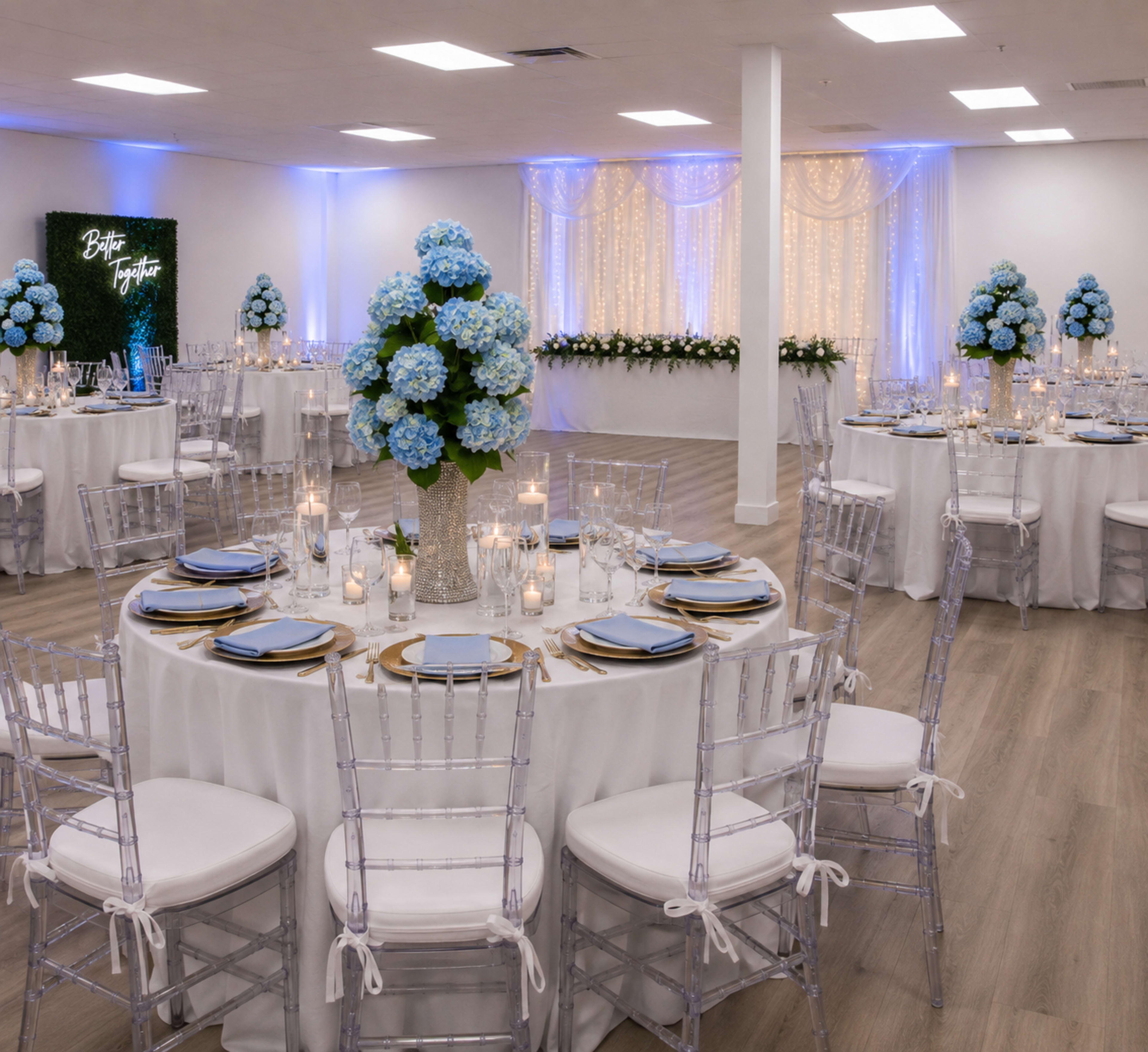 The image shows a decorated event space with round tables adorned with light blue floral centerpieces and white tablecloths, illuminated by soft blue lighting and surrounded by elegant Chiavari chairs.