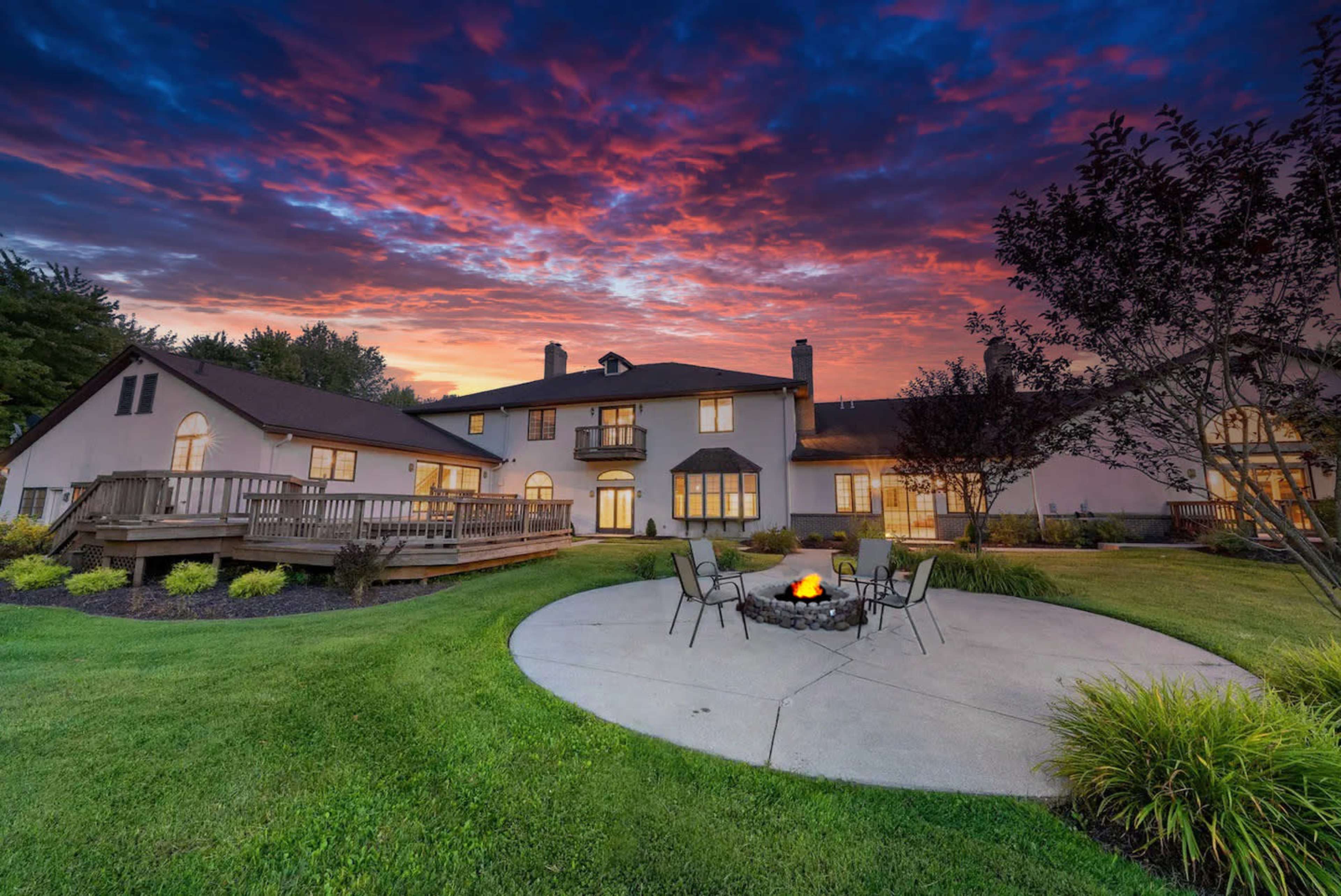 A large house is set against a vibrant sunset, with a circular patio featuring a fire pit and chairs in the foreground.