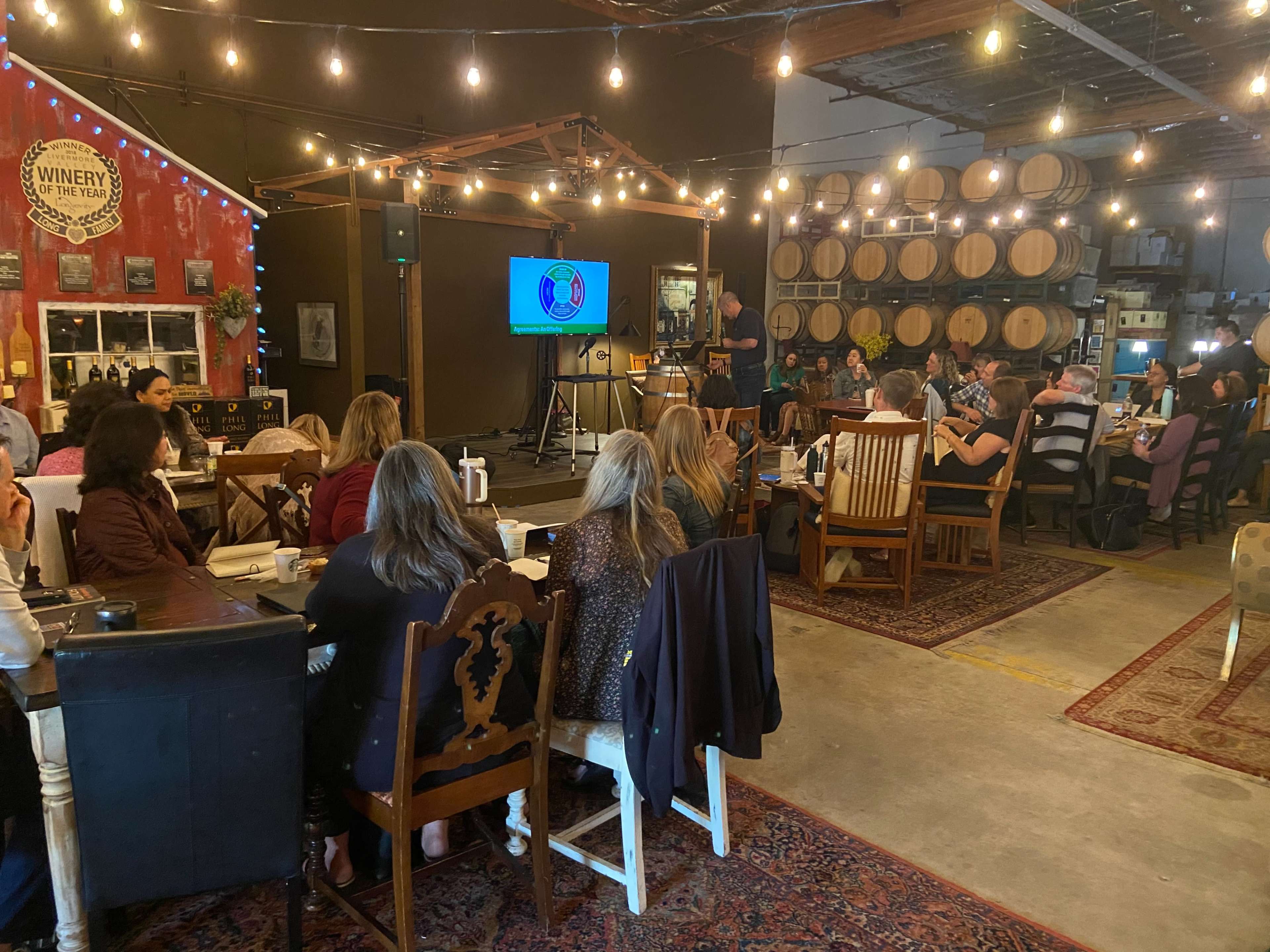 A gathering of people is taking place in a winery, with barrels in the background and a screen displaying a presentation.