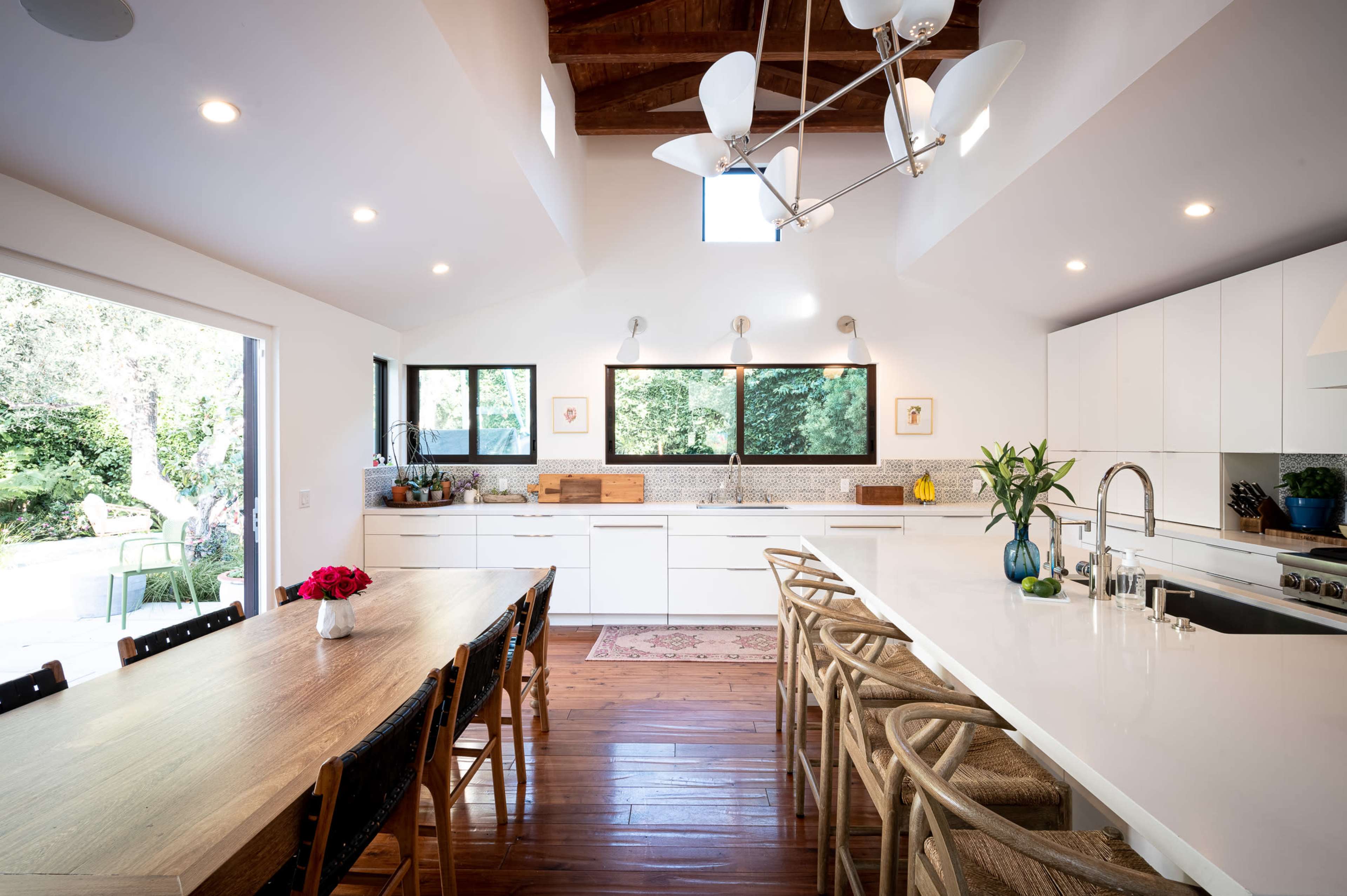 A modern kitchen features white cabinets, a large island with seating, and a dining table, with large windows allowing natural light to illuminate the space.