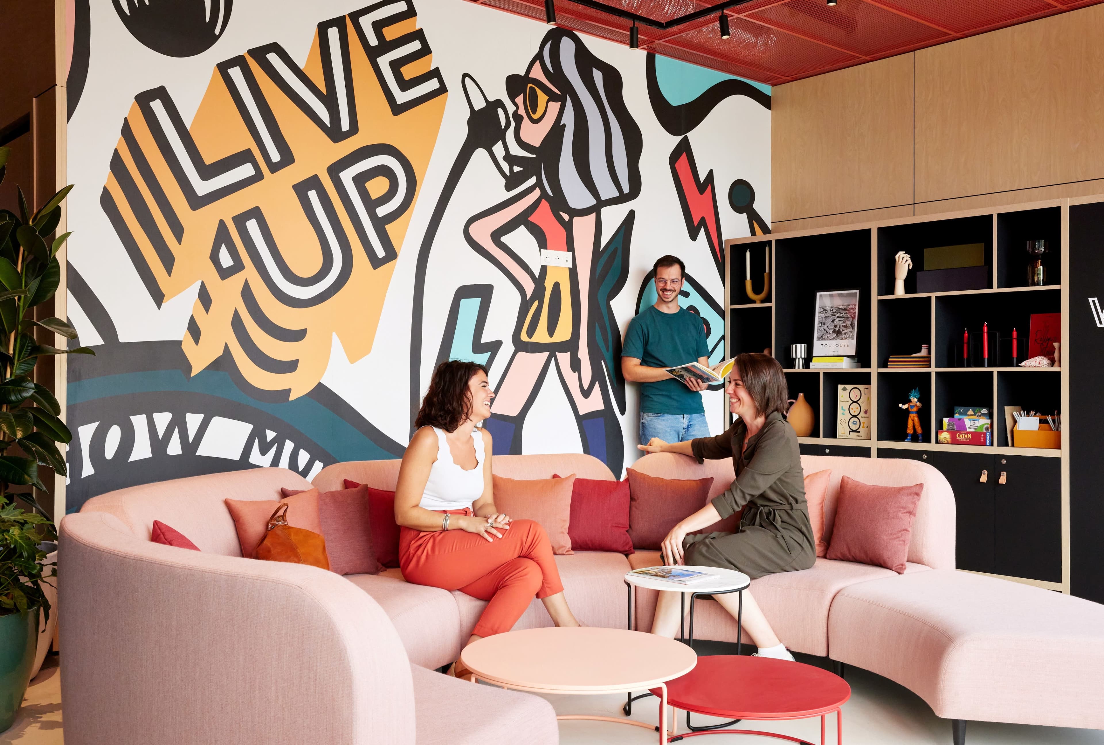 Three people are seated in a modern lounge area with a large, colorful mural on the wall that features graphic designs and text.