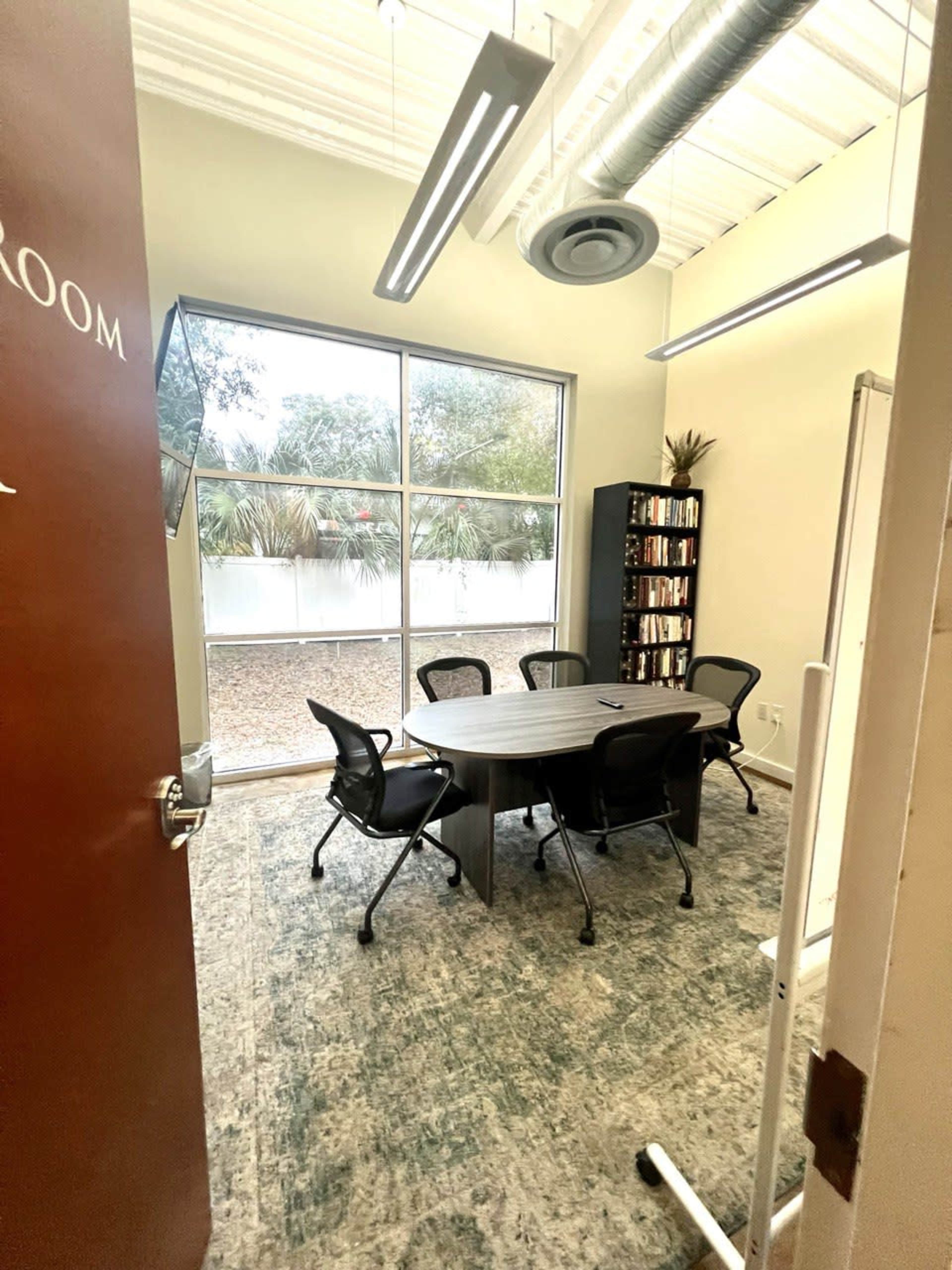 An office conference room with a round table and chairs, illuminated by overhead lighting and featuring large windows.