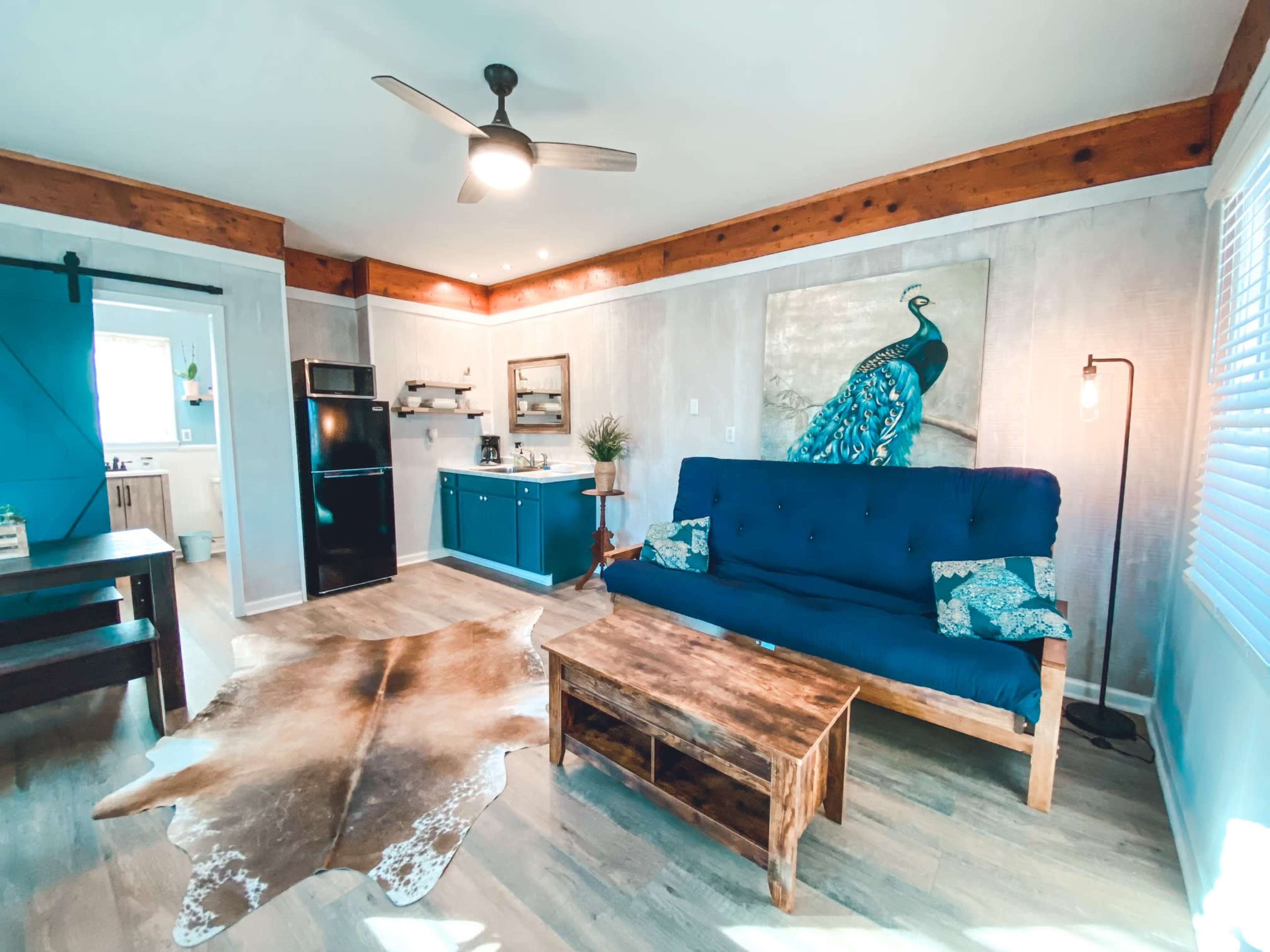 A cozy living room featuring a blue sofa, a wooden coffee table, and a peacock painting on the wall, with a kitchen area visible in the background.