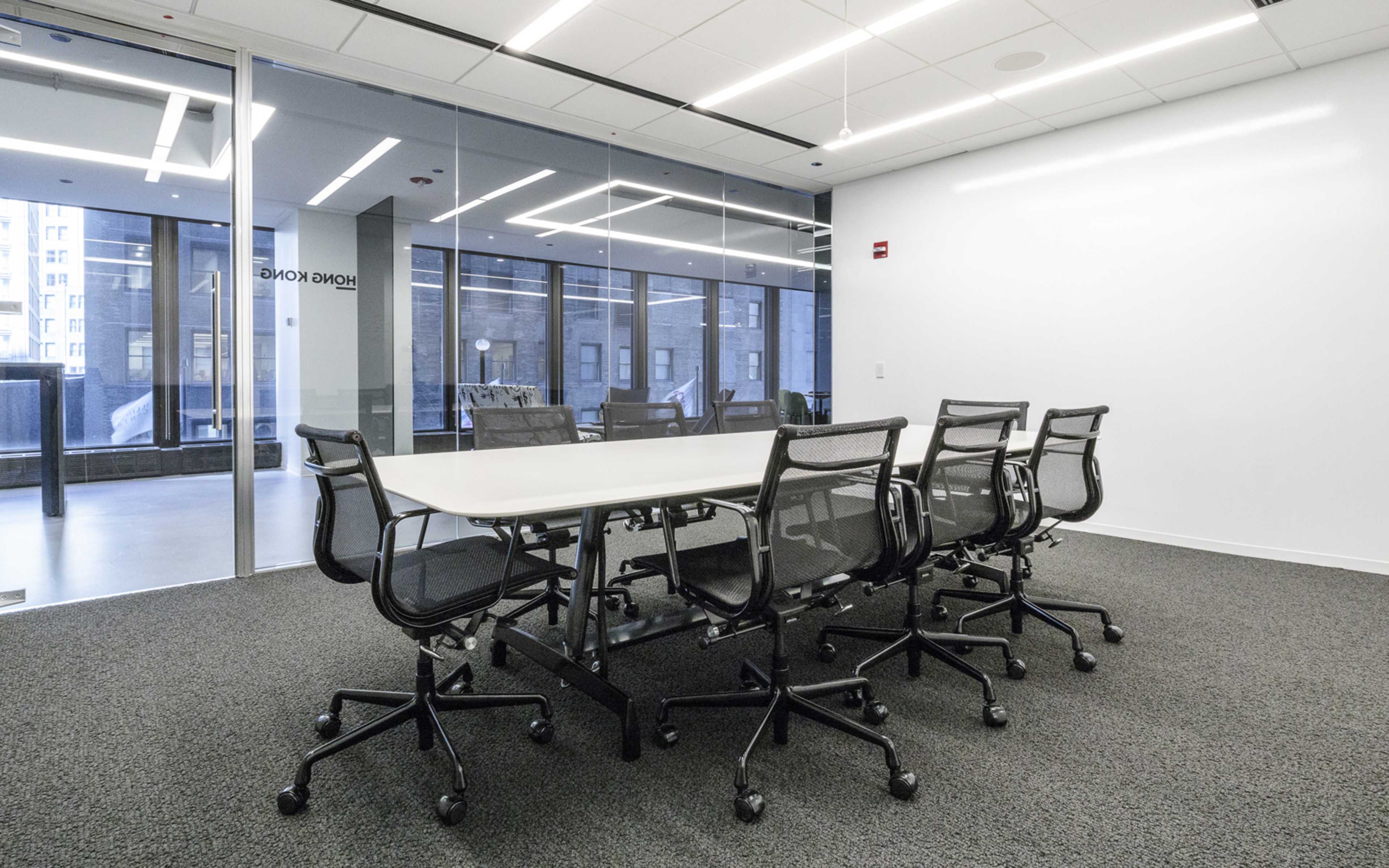 A modern conference room features a large, rectangular table surrounded by six black mesh chairs, with glass walls offering a view of an urban landscape.