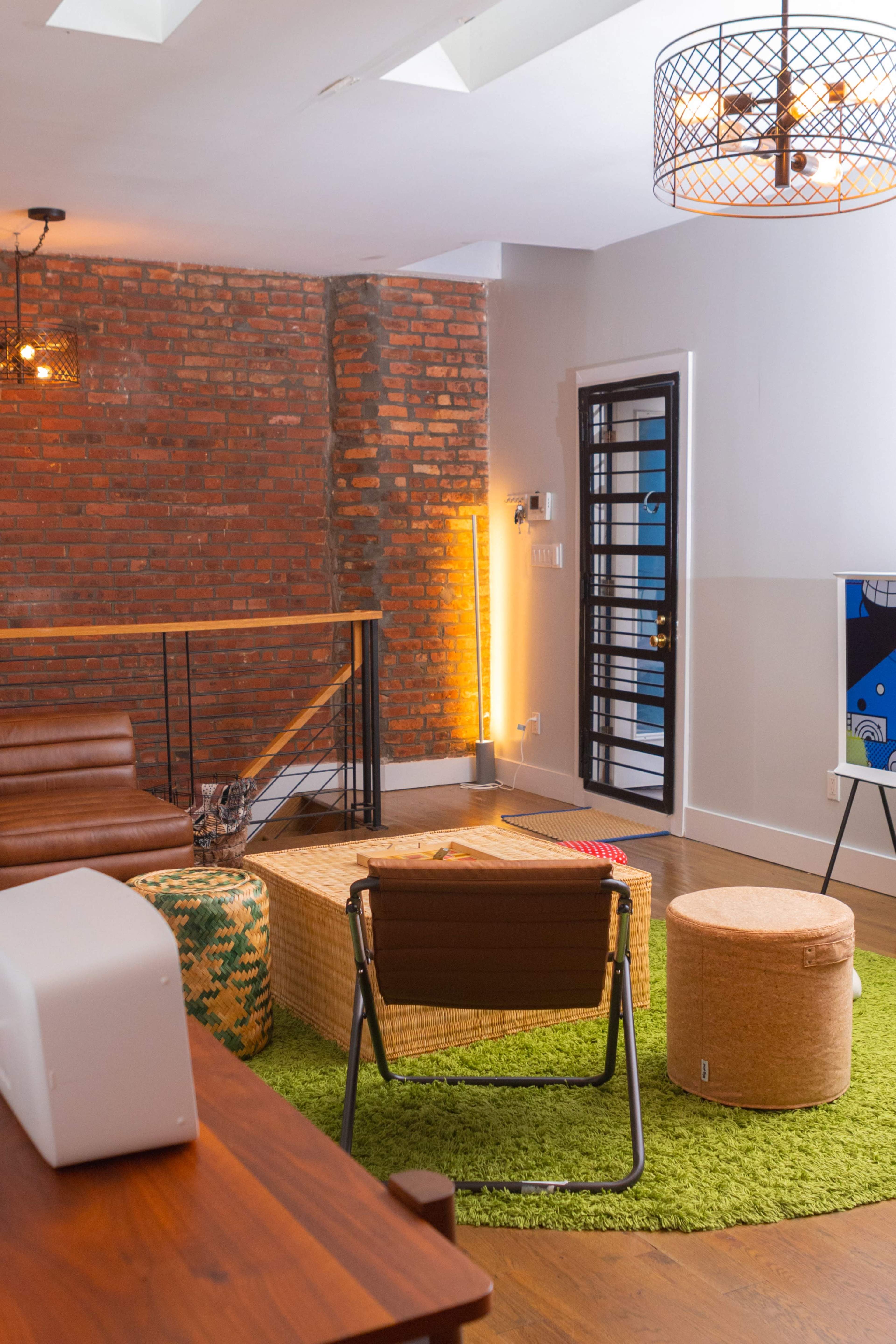 A modern lounge area featuring exposed brick walls, a brown leather sofa, various seating options, and a green rug.