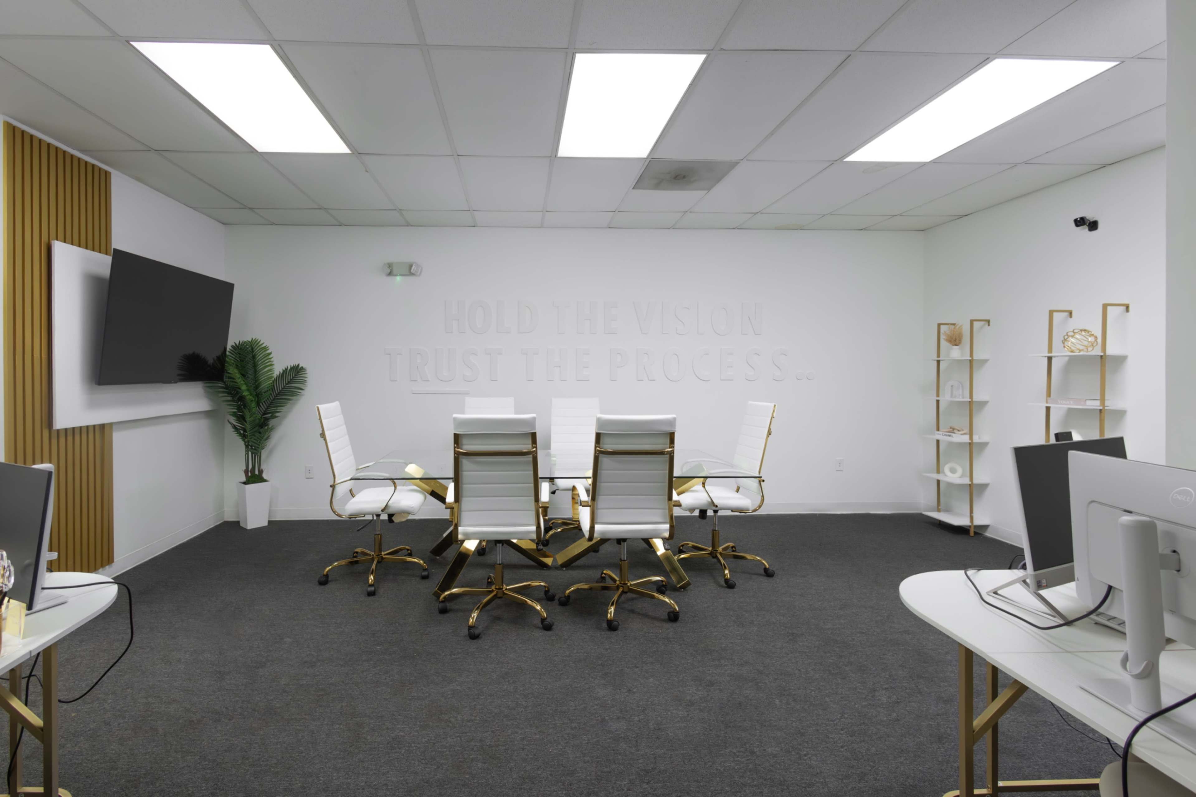 A modern conference room features a round table surrounded by white chairs, a large screen on the wall, and minimalist decor.