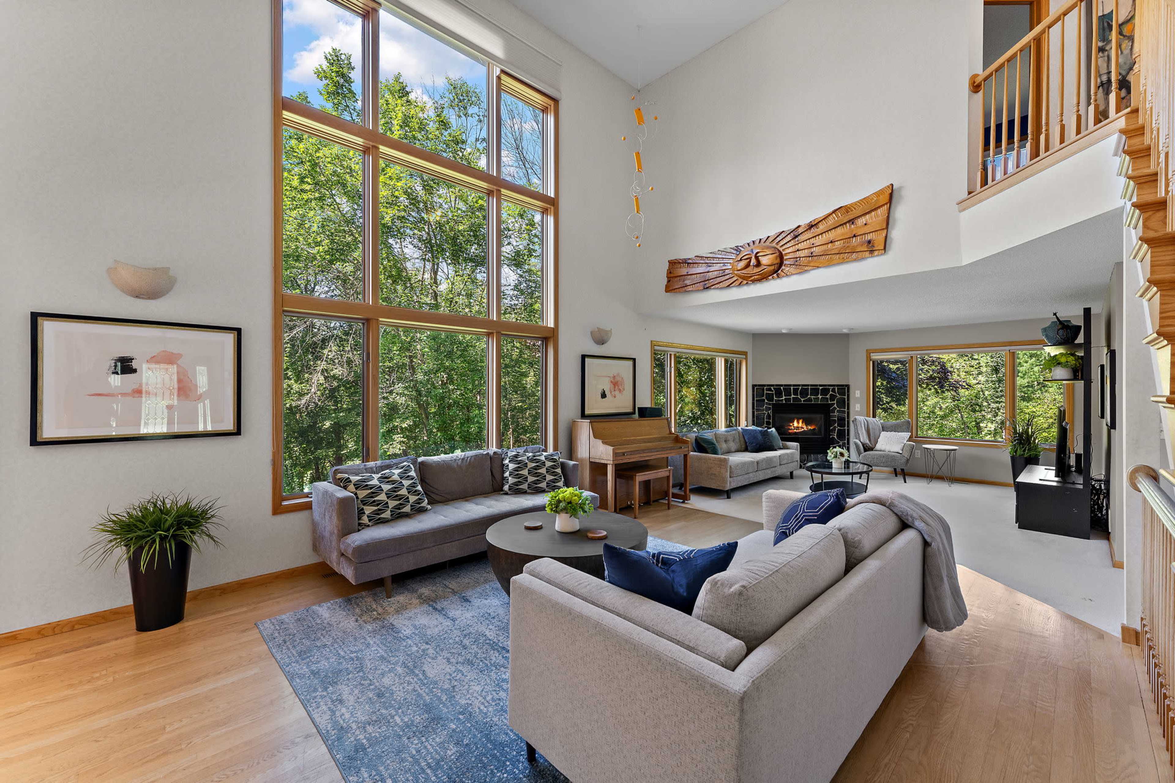 The image shows a spacious living room with large windows, two sofas, a coffee table, and a fireplace, surrounded by greenery outside.