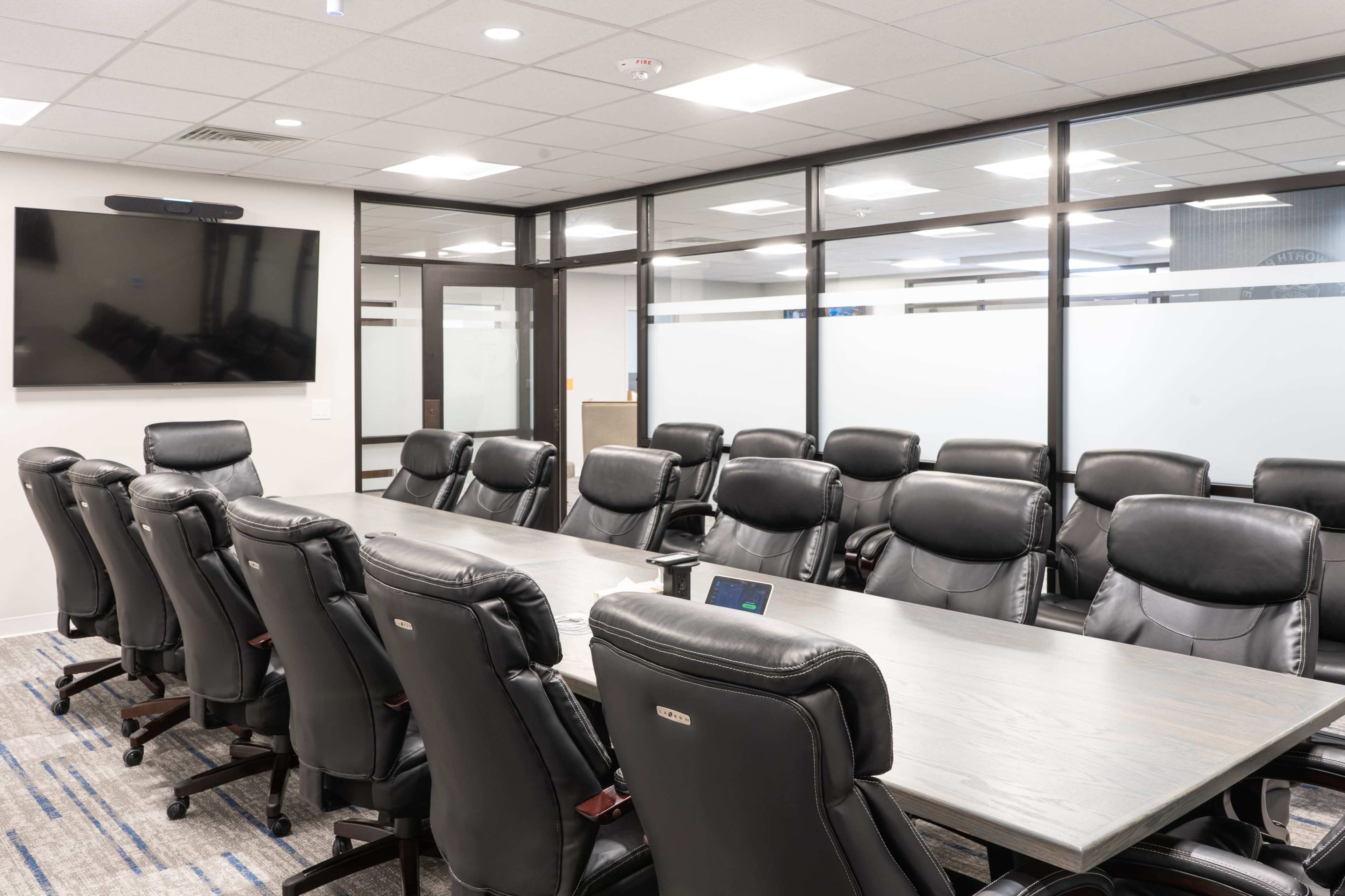 A conference room featuring a long wooden table surrounded by black leather chairs and a flat-screen television mounted on the wall.