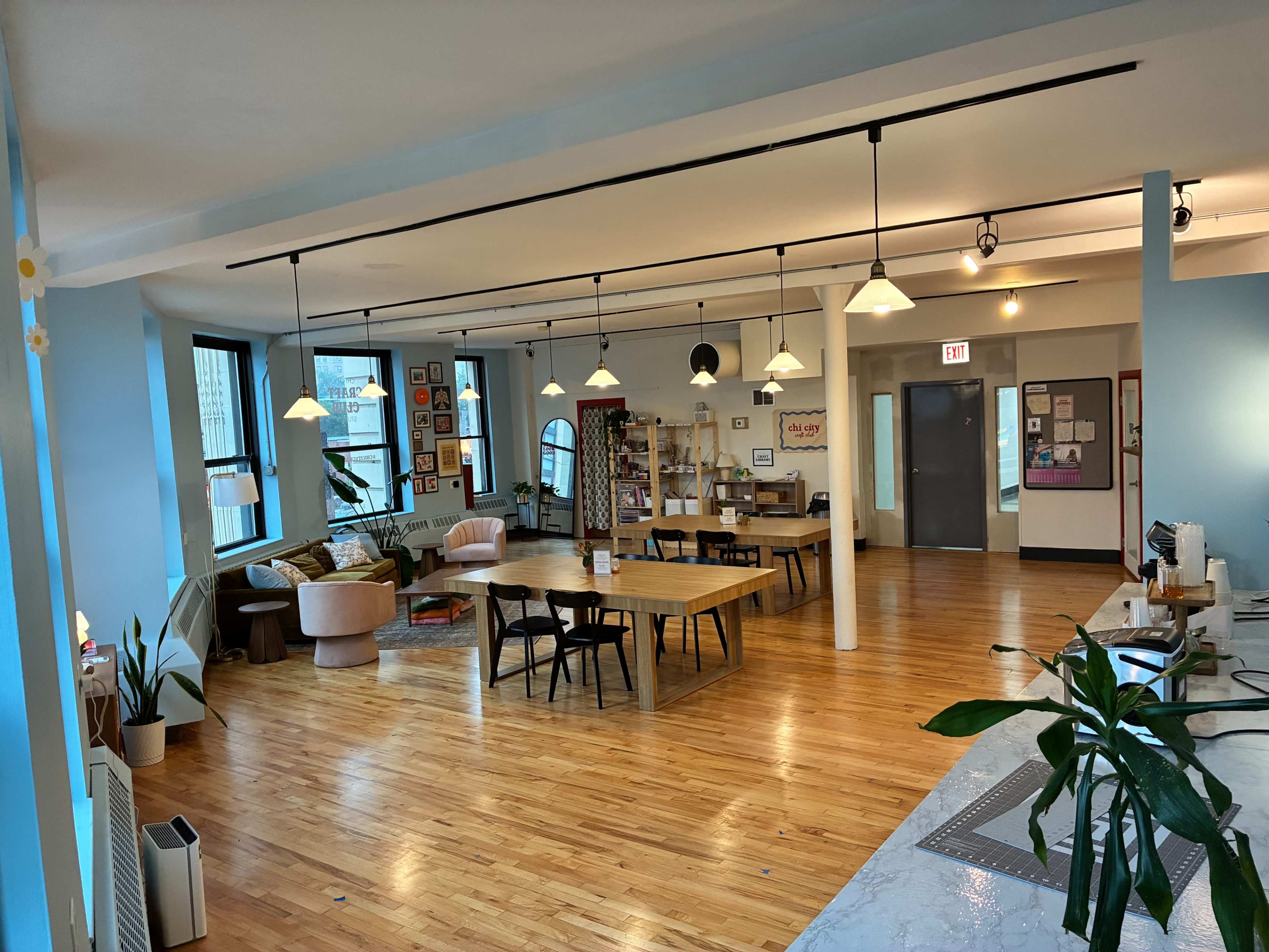 The image shows a spacious, well-lit communal workspace featuring wooden floors, large windows, various seating arrangements, and decorative plants.