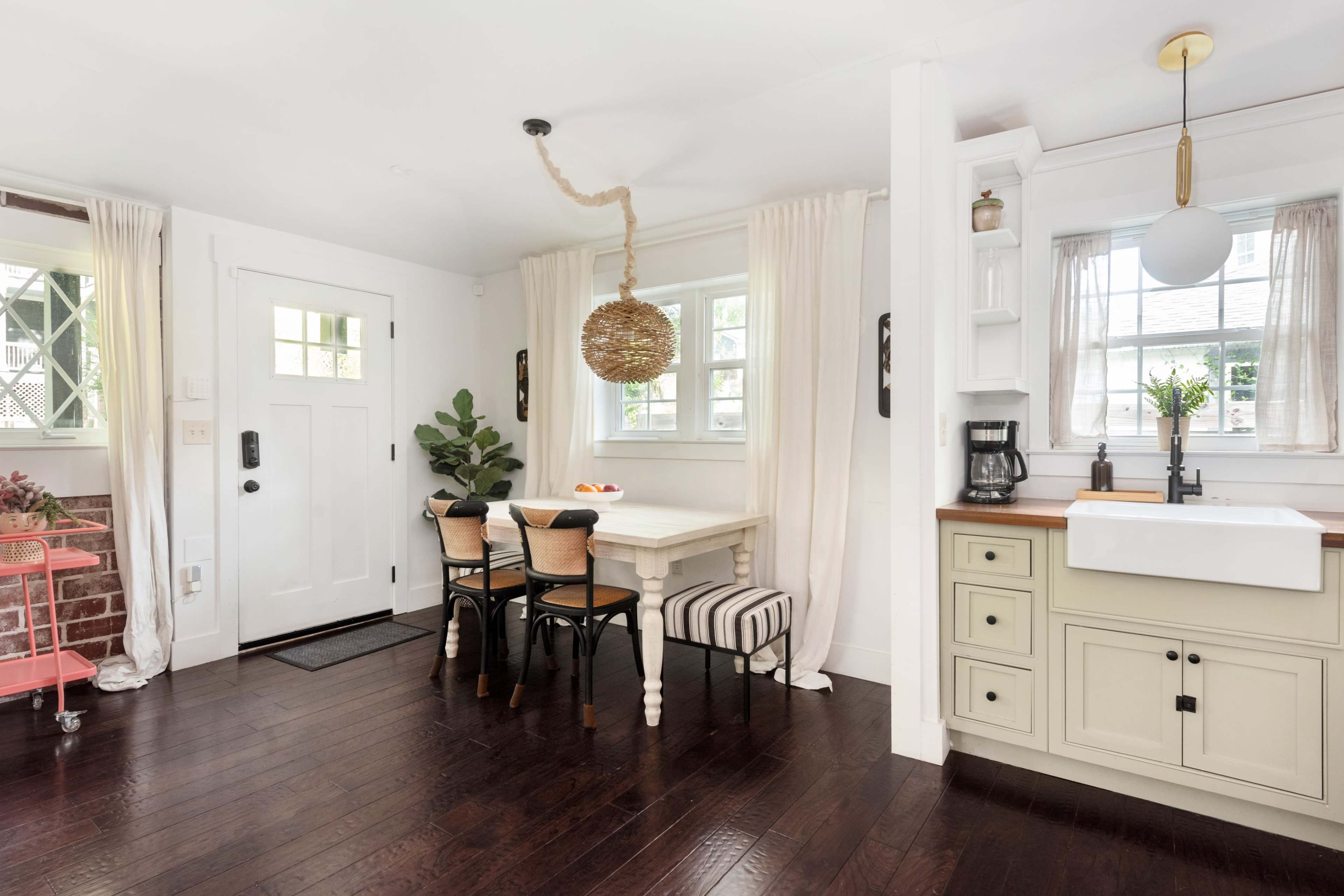 A bright, modern kitchen features a small dining table with two chairs, a sink area, and a decorative pendant light, complemented by white walls and rich wooden flooring.