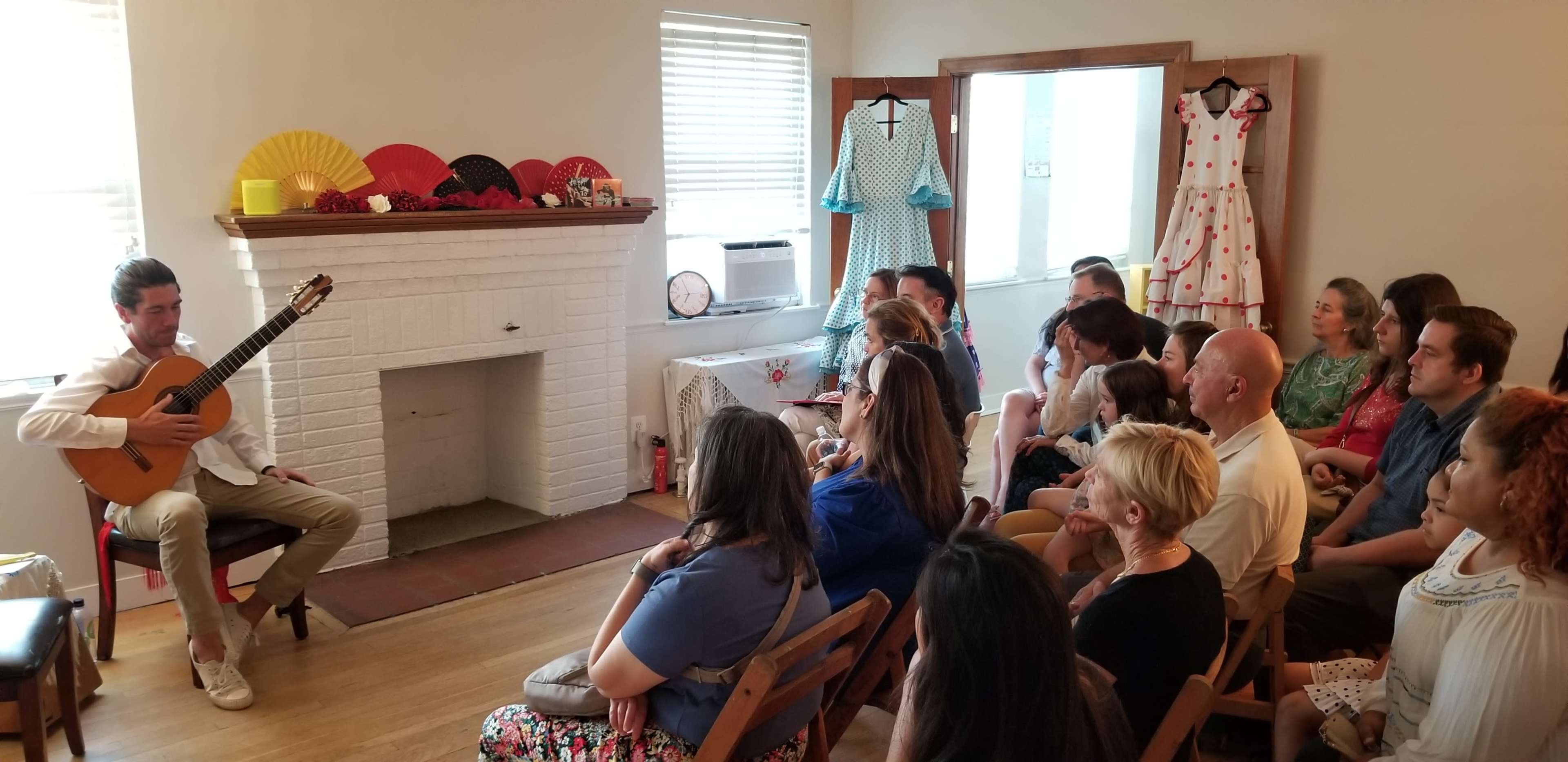 A musician plays guitar for an audience seated in a room decorated with clothing and fans.