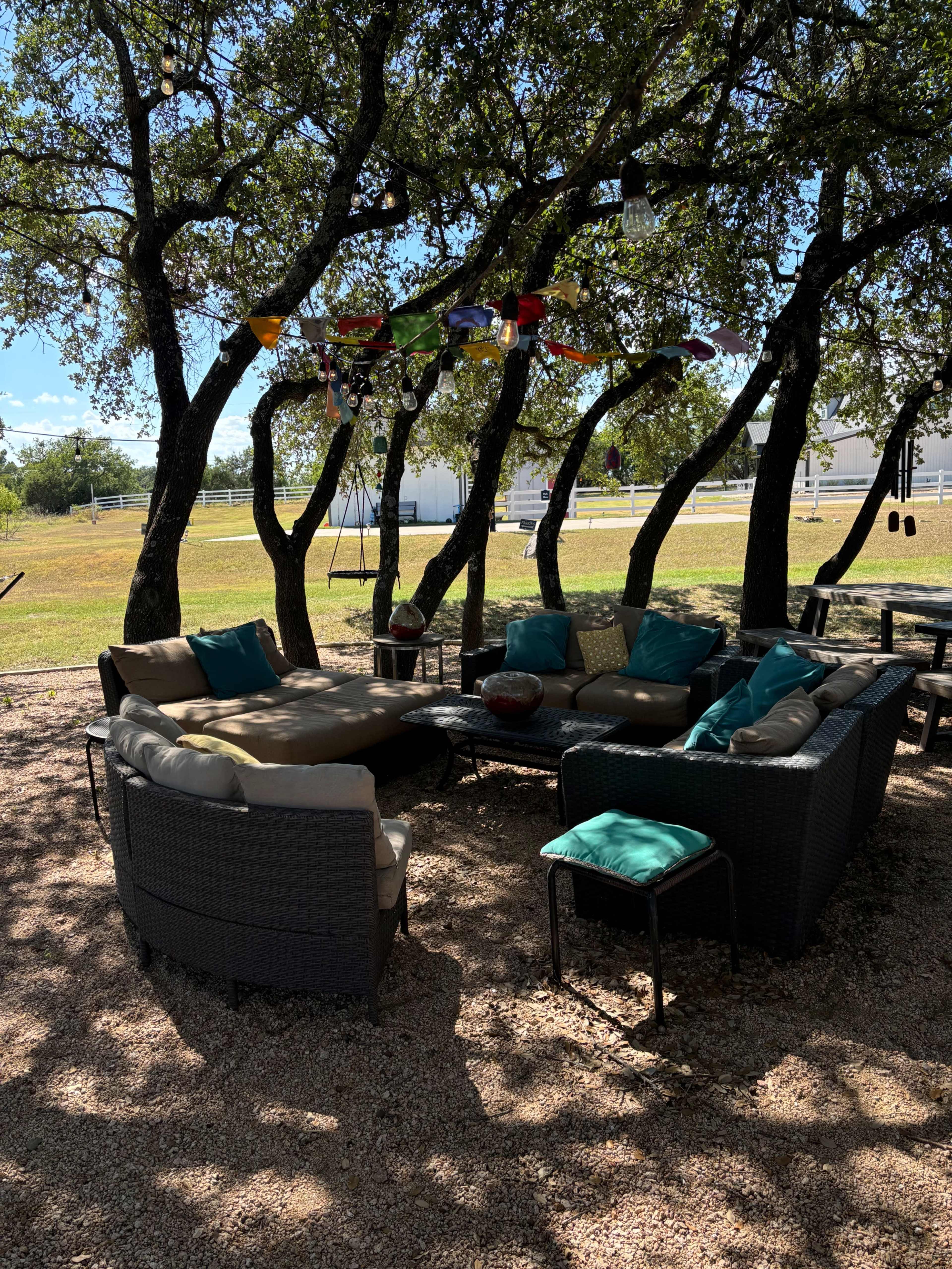 A circular seating area with cushions is set under a group of trees, surrounded by gravel and decorated with hanging items.