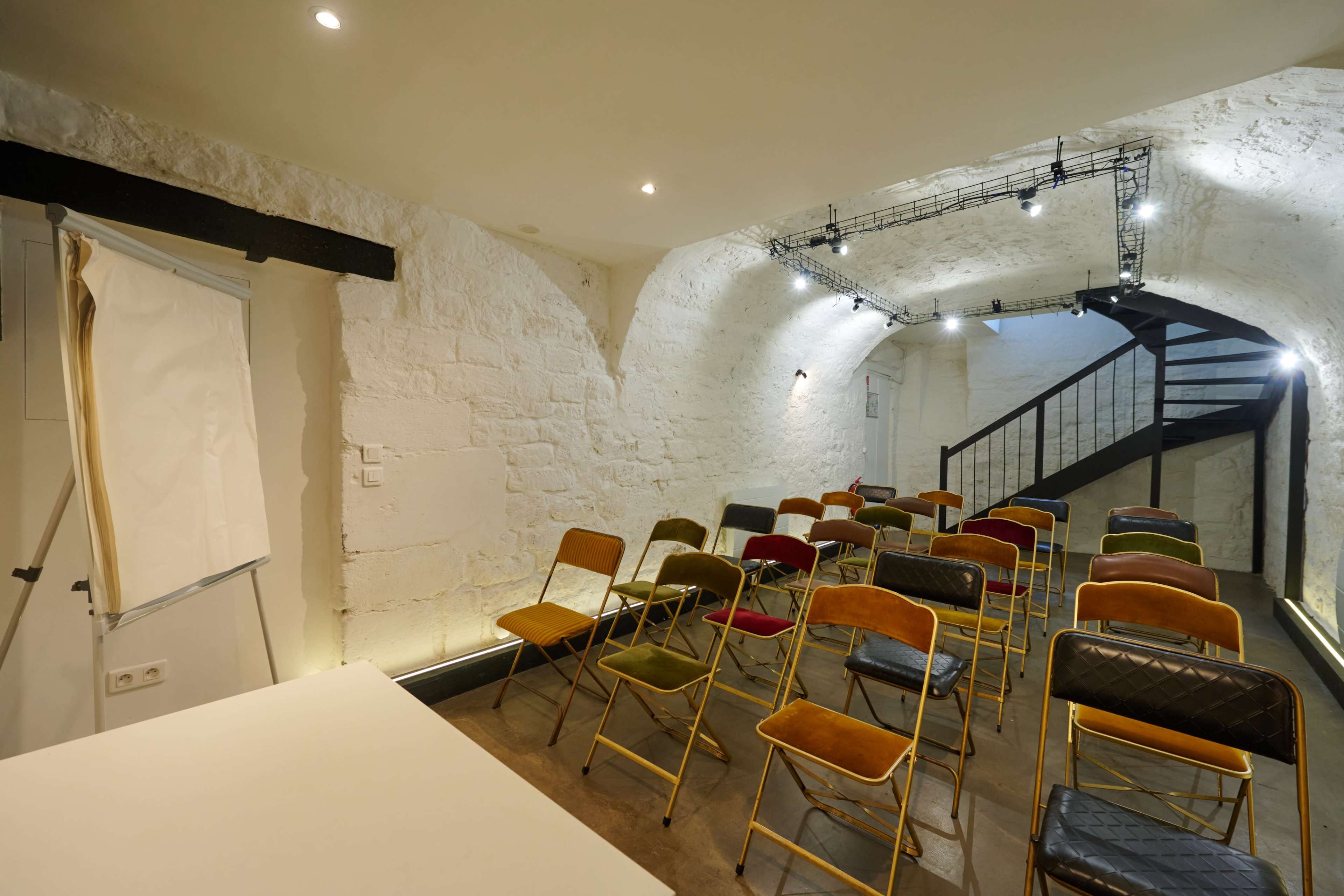 A basement room arranged with rows of colorful folding chairs facing a blank projection screen and a staircase in the corner.