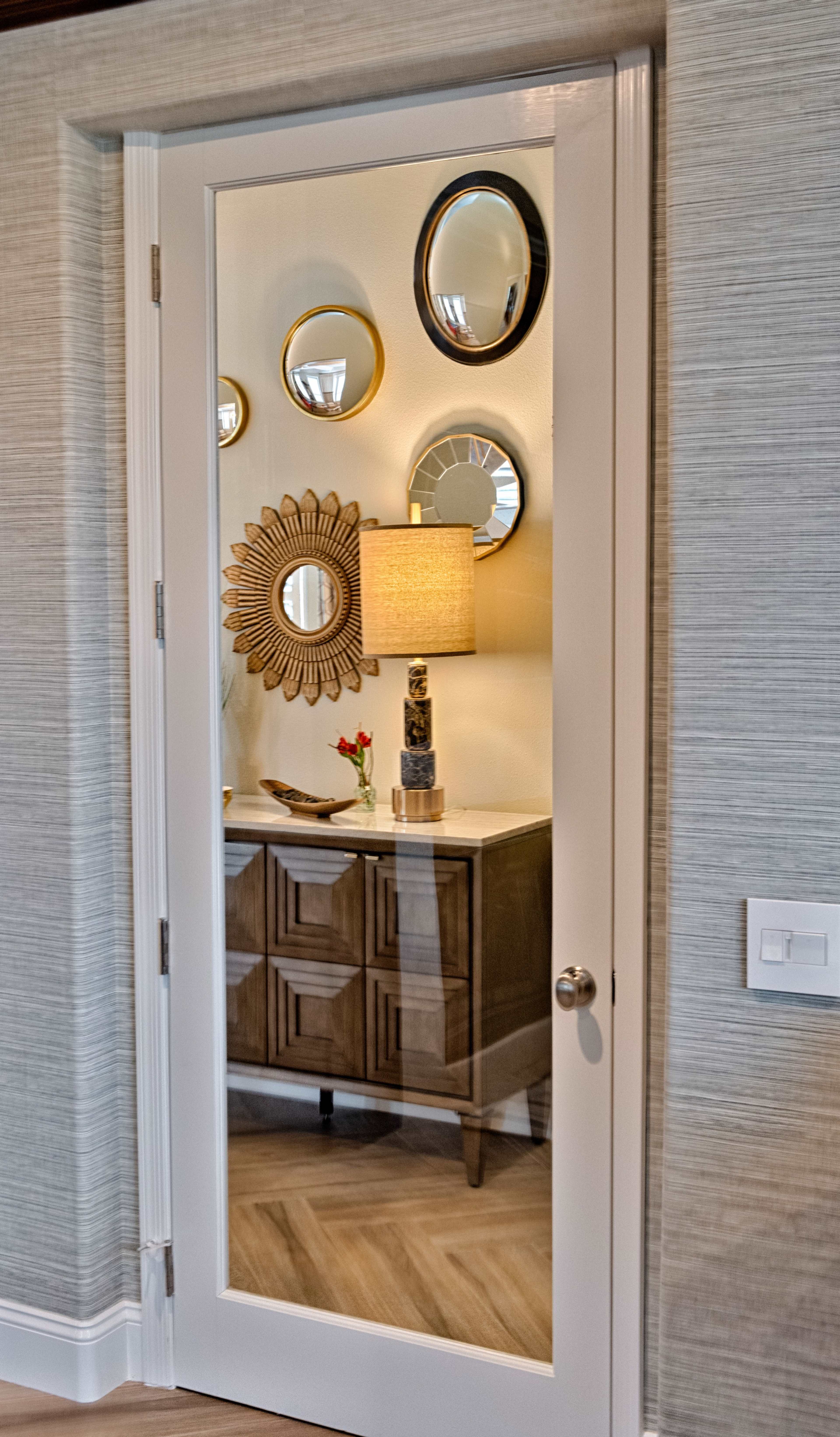 A mirror-paneled door reflects a console table adorned with a lamp and decorative mirrors against a textured wall.