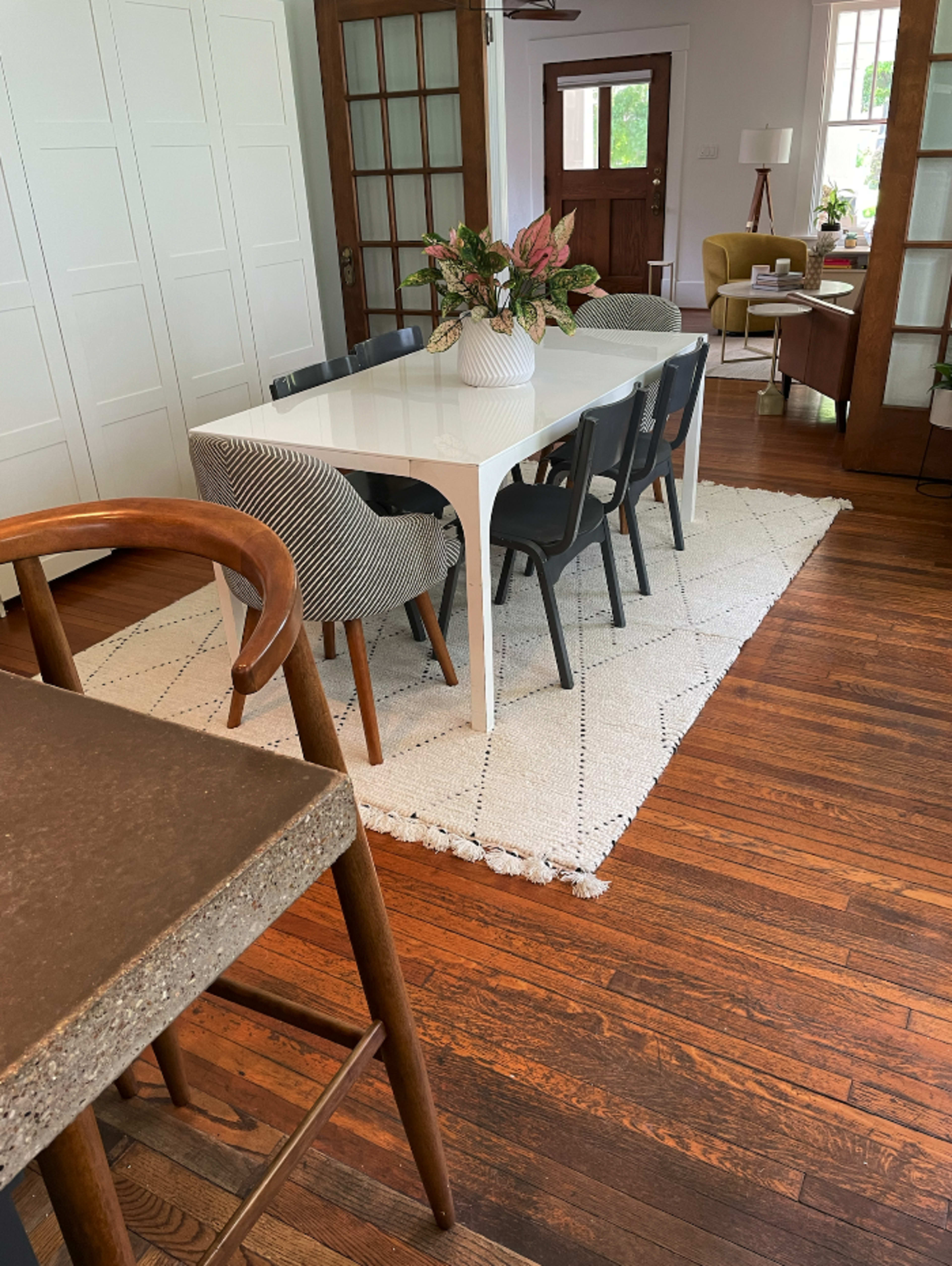 A modern dining area features a white table surrounded by six chairs, with a decorative rug beneath and a plant centerpiece on the table.